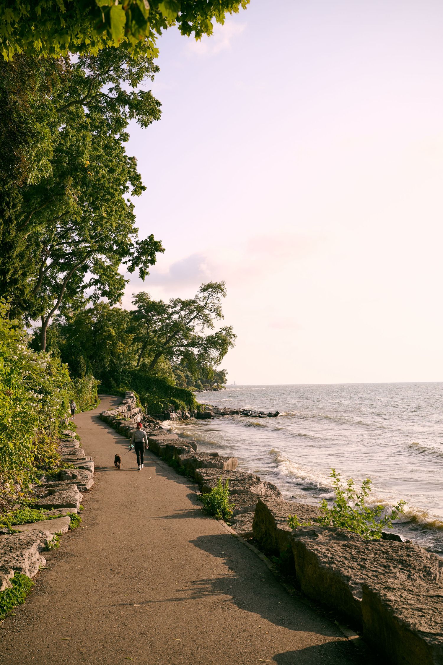 A tranquil coastal path winds along rocky cliffs with green trees and an ocean view on a cloudy day.