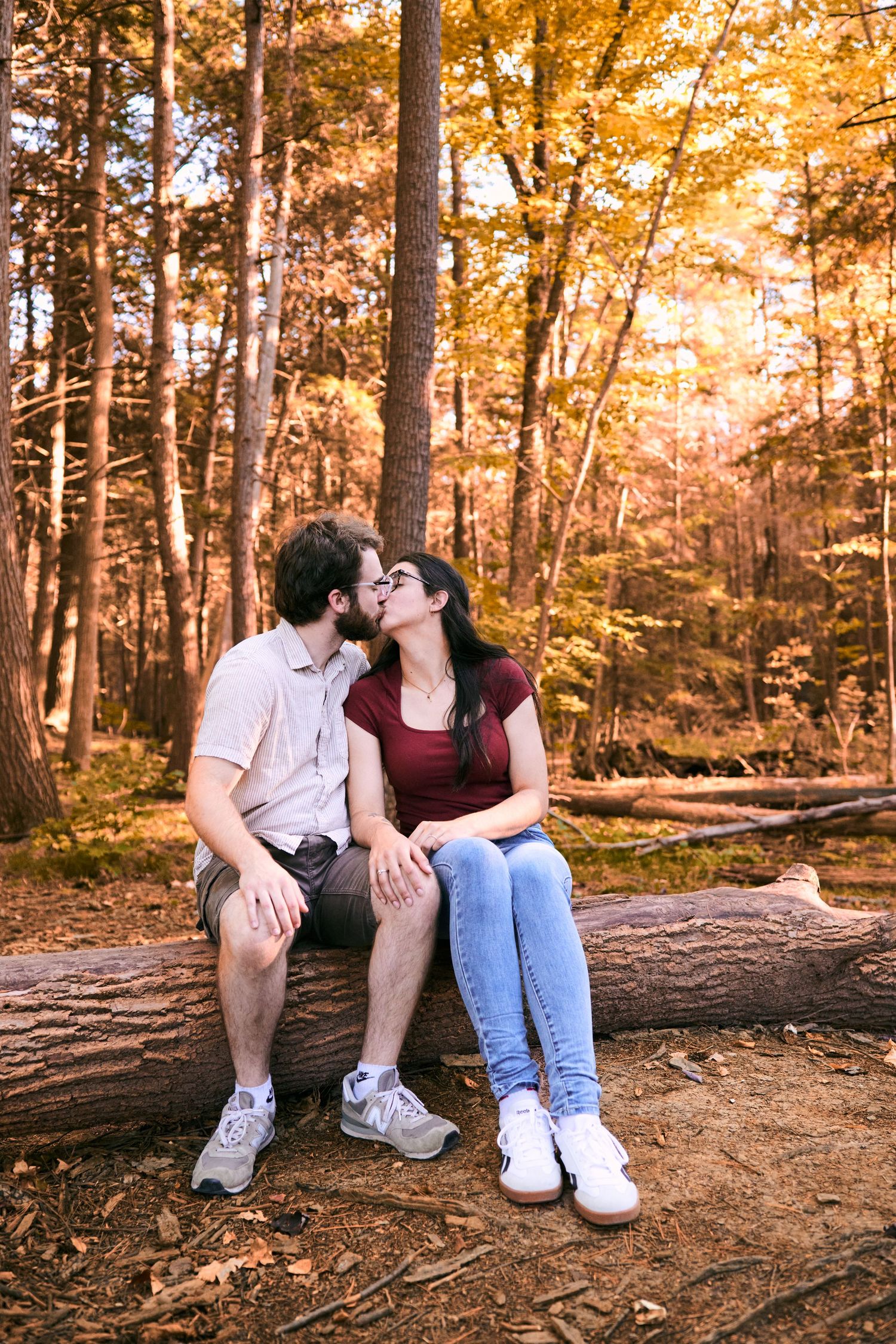 A couple sits together on a fallen log in an autumn forest bathed in warm golden sunlight.