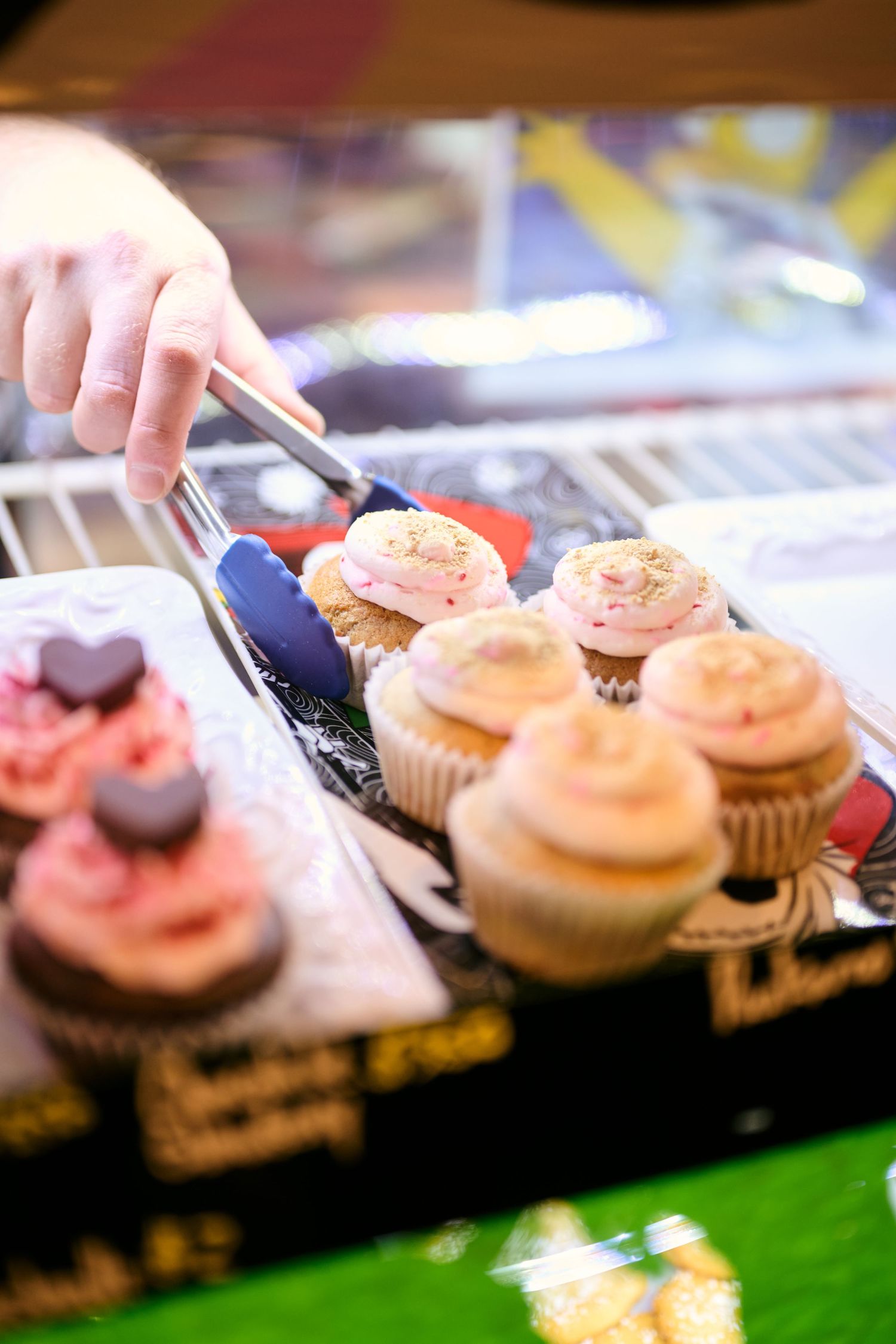 A hand selecting colorful frosted cupcakes from a bakery display case with tongs.