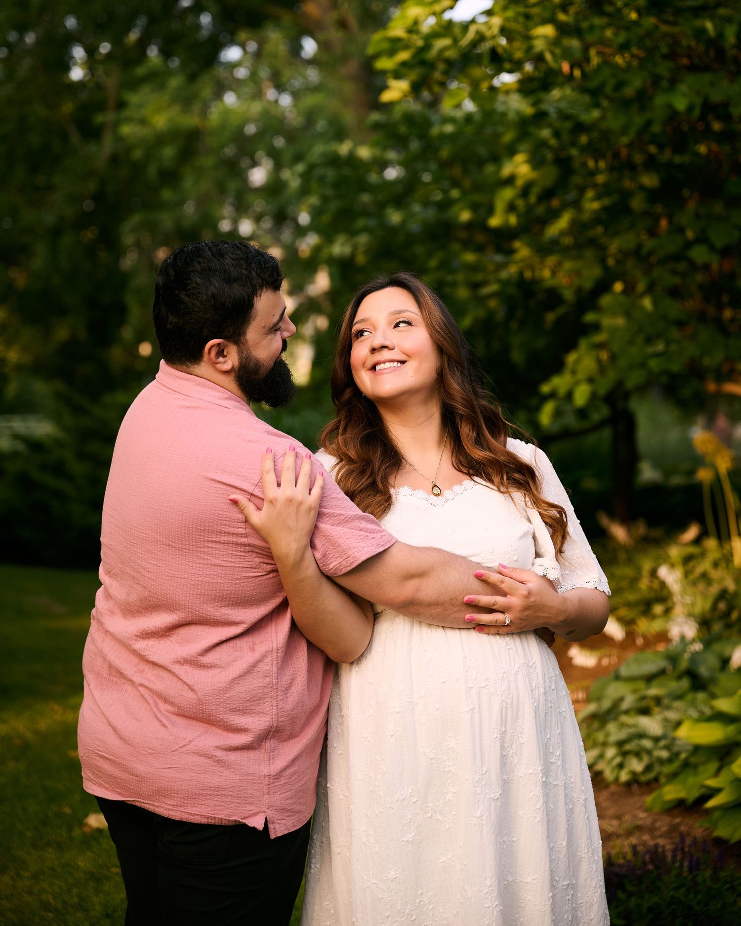 A couple in matching pink and white outfits share a tender moment in a lush garden, the woman cradling her baby bump.