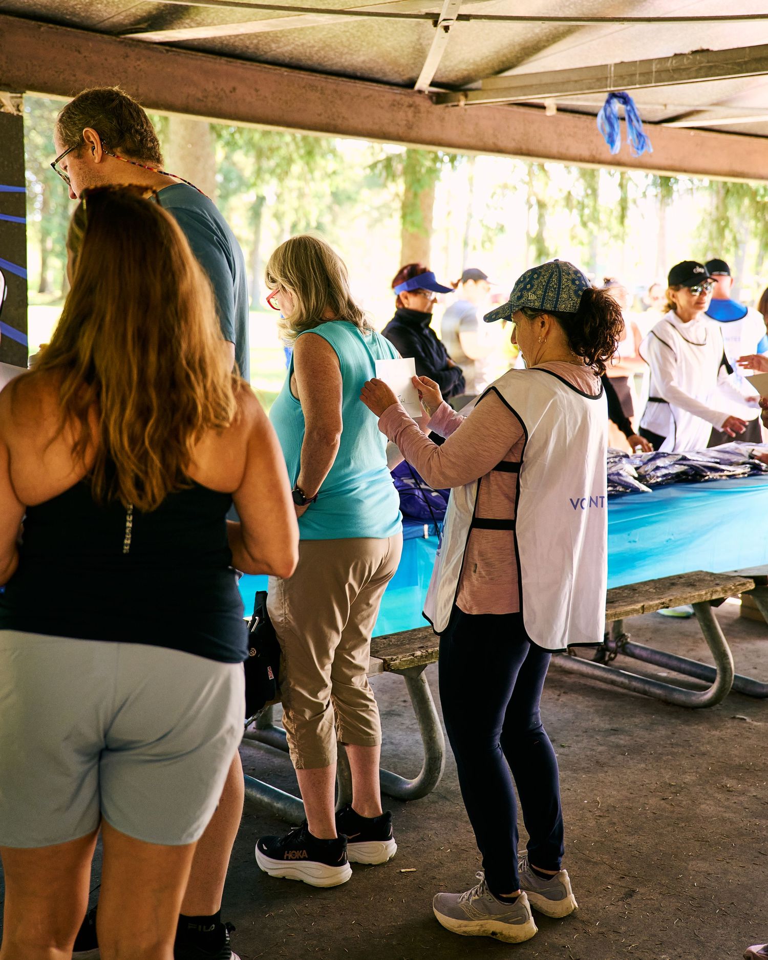 People gathered under a shaded pavilion at an outdoor event, browsing items on a blue table.