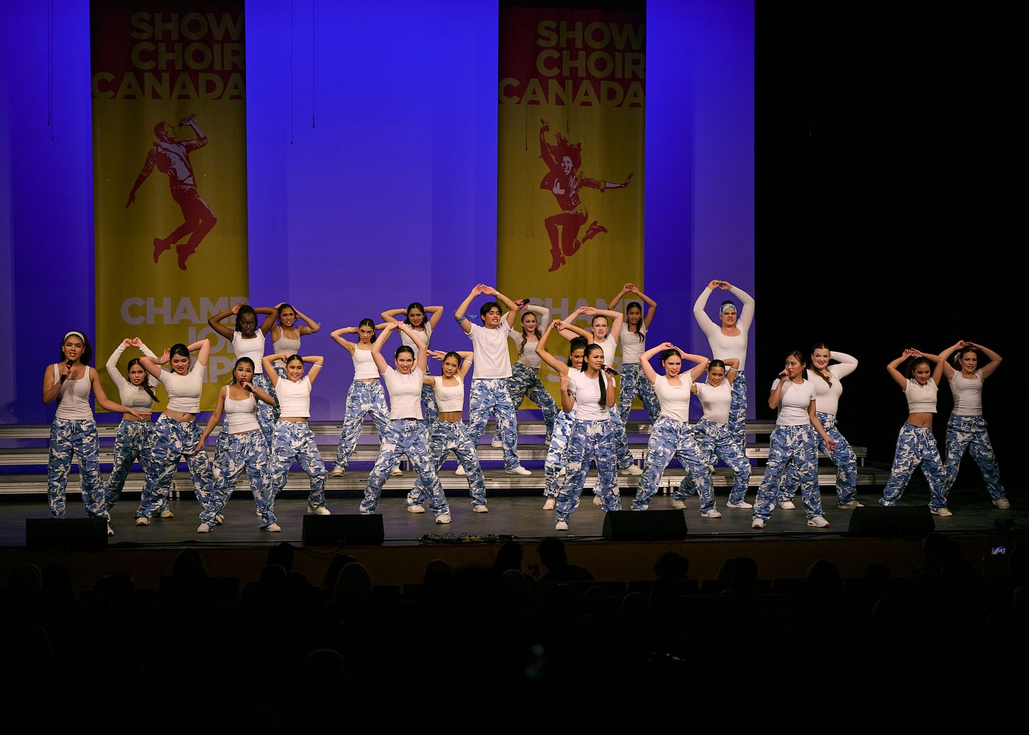 A large show choir group performs a synchronized dance routine in matching blue camo pants on a competition stage.