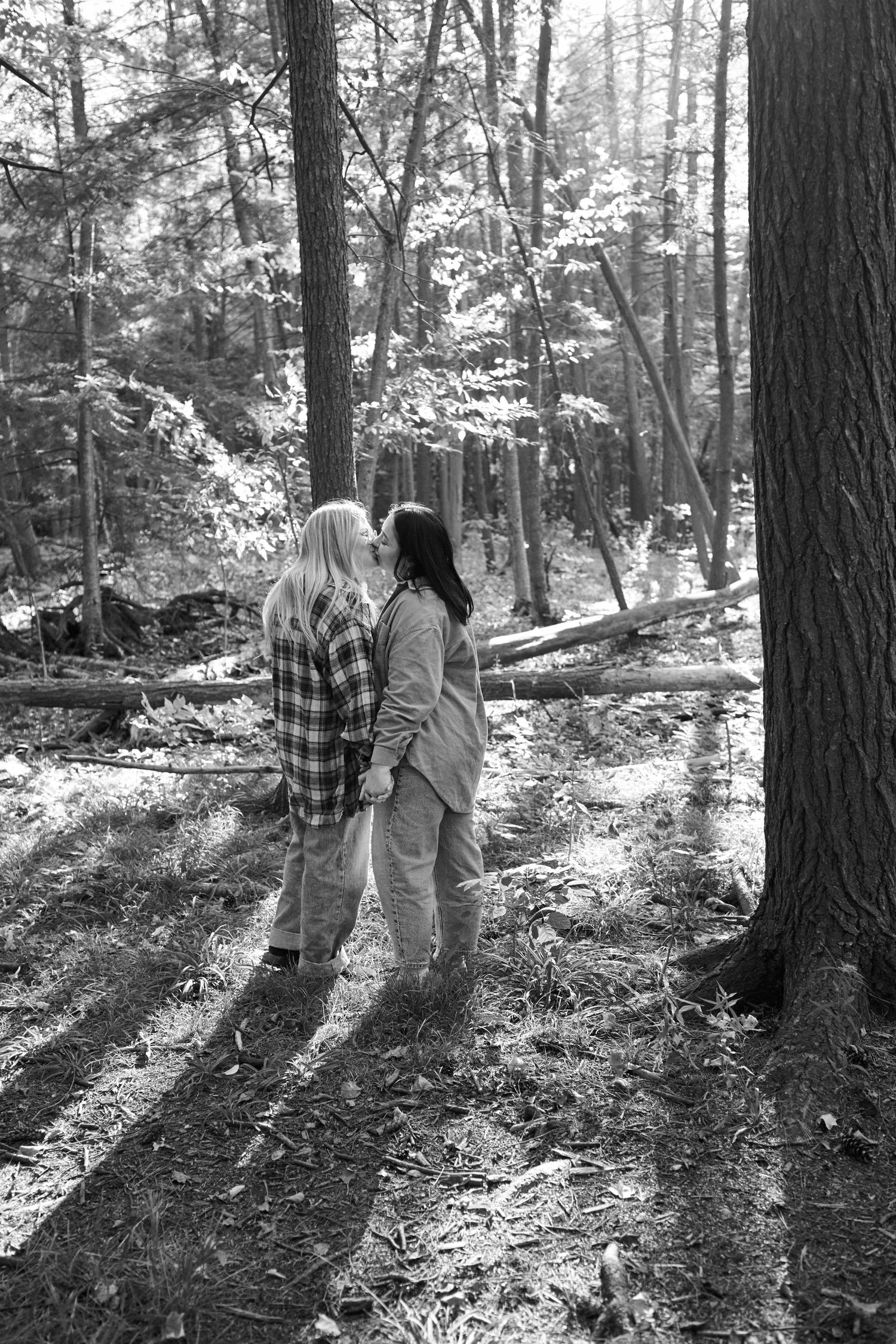 A couple in plaid and white outfits walks together through a sunlit forest with tall pine trees.