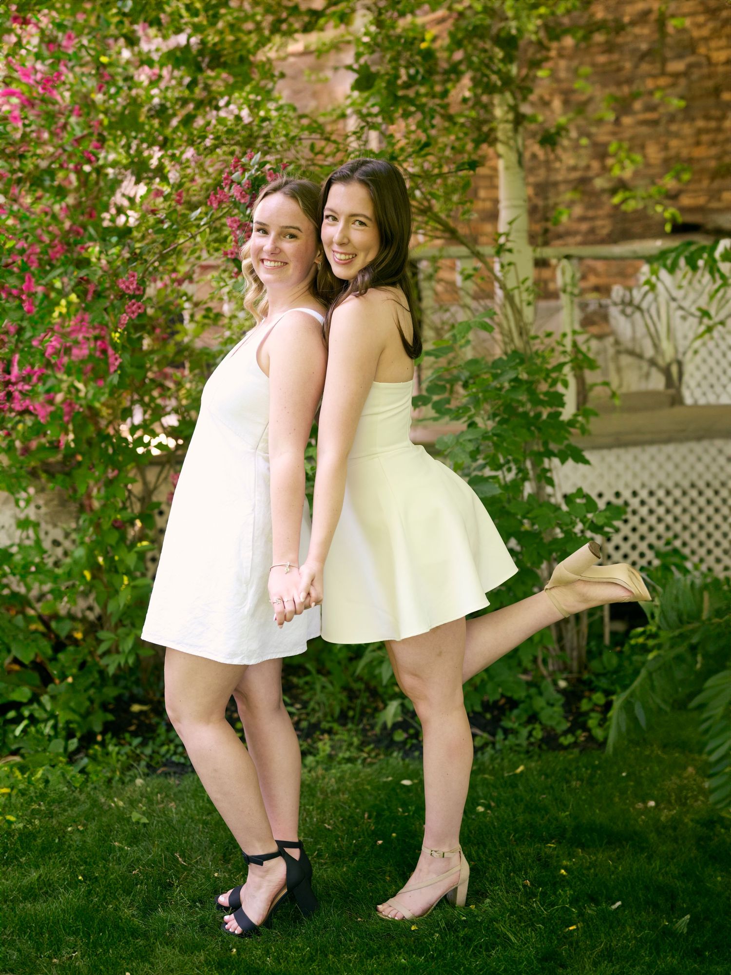 Two people in matching white and cream dresses pose together in a garden setting with pink flowering bushes.