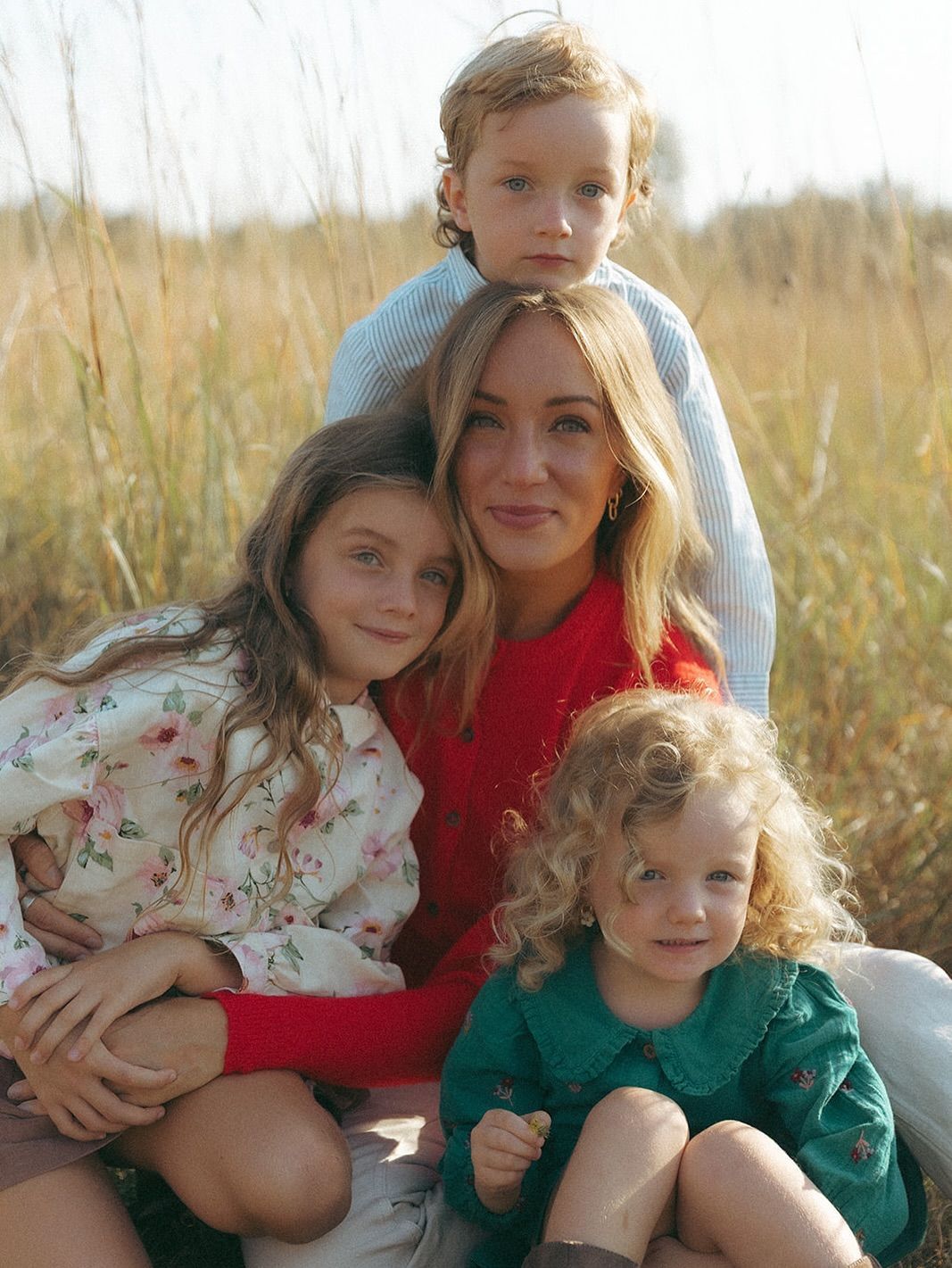 A woman and three young children pose together in a golden field, wearing colorful autumn outfits.