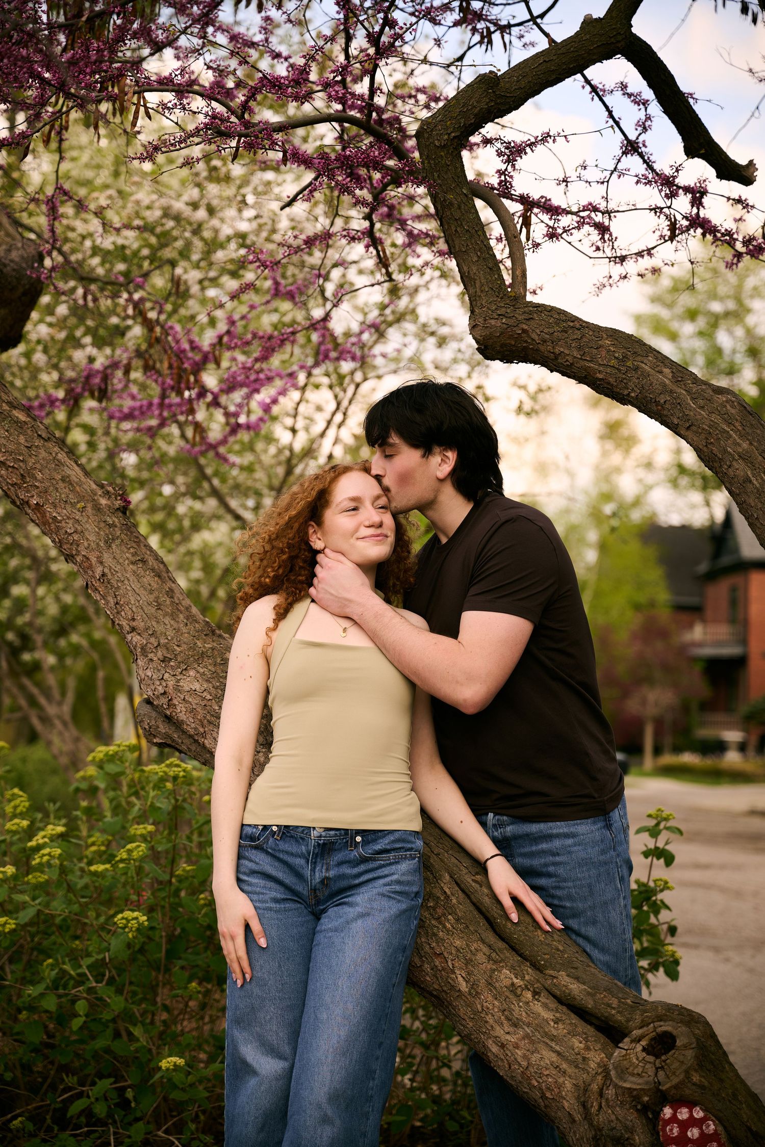 A couple shares a tender kiss under blooming spring trees, with the man gently holding the woman's face outdoors.