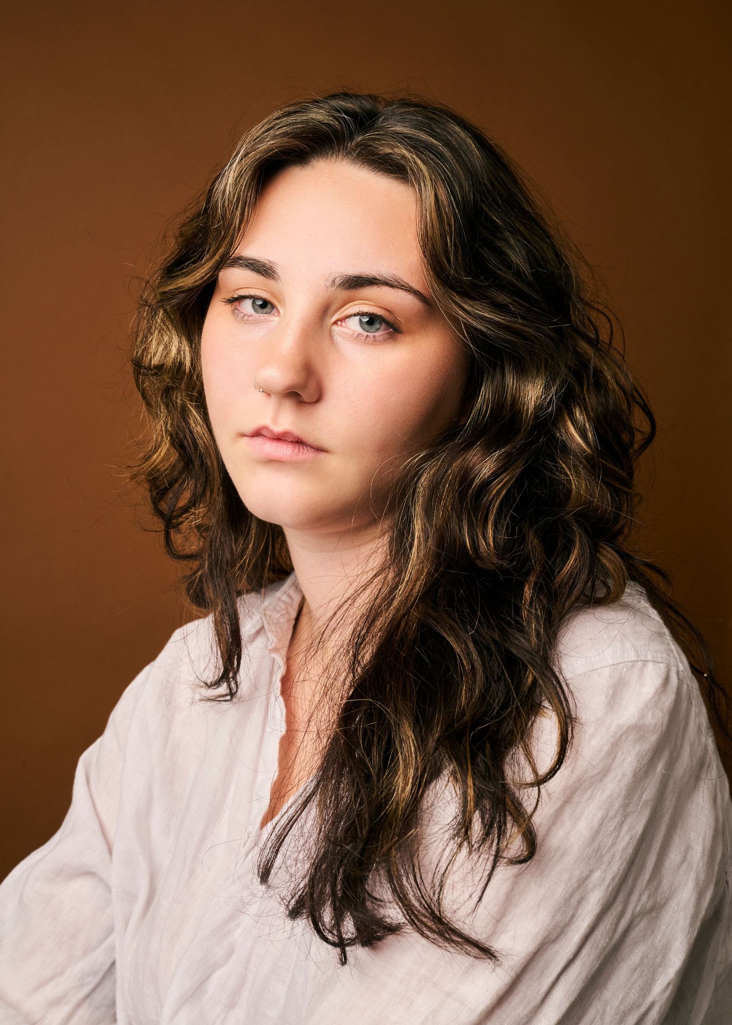 Studio portrait series showing a contemplative subject with wavy dark hair wearing a relaxed white button-down shirt against brown backdrop.