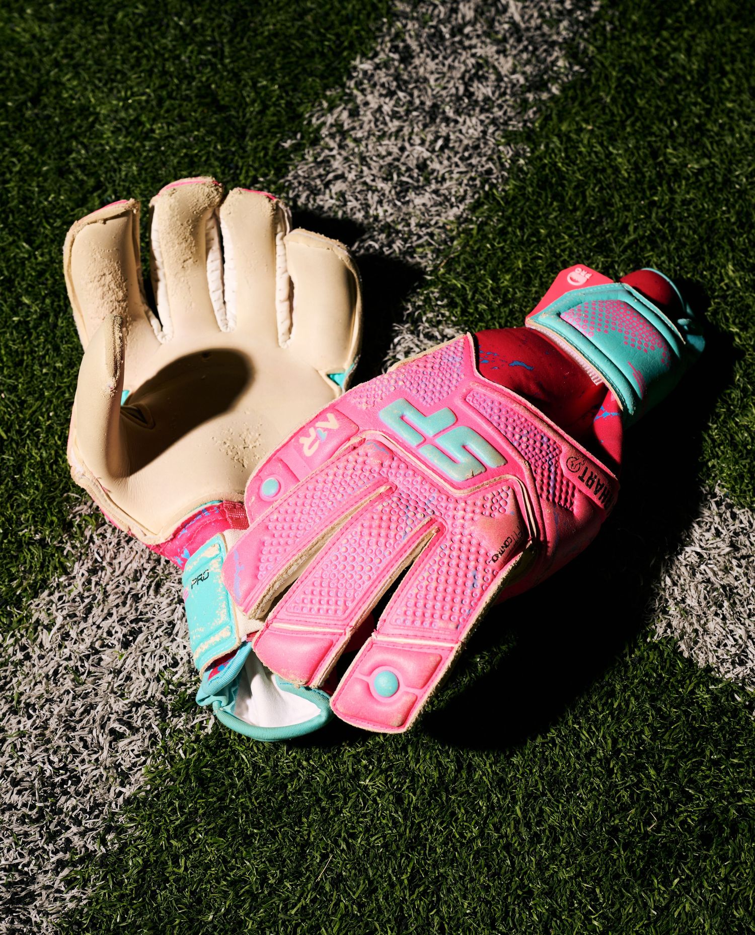 Pink and turquoise softball batting gloves on artificial turf with white chalk line.