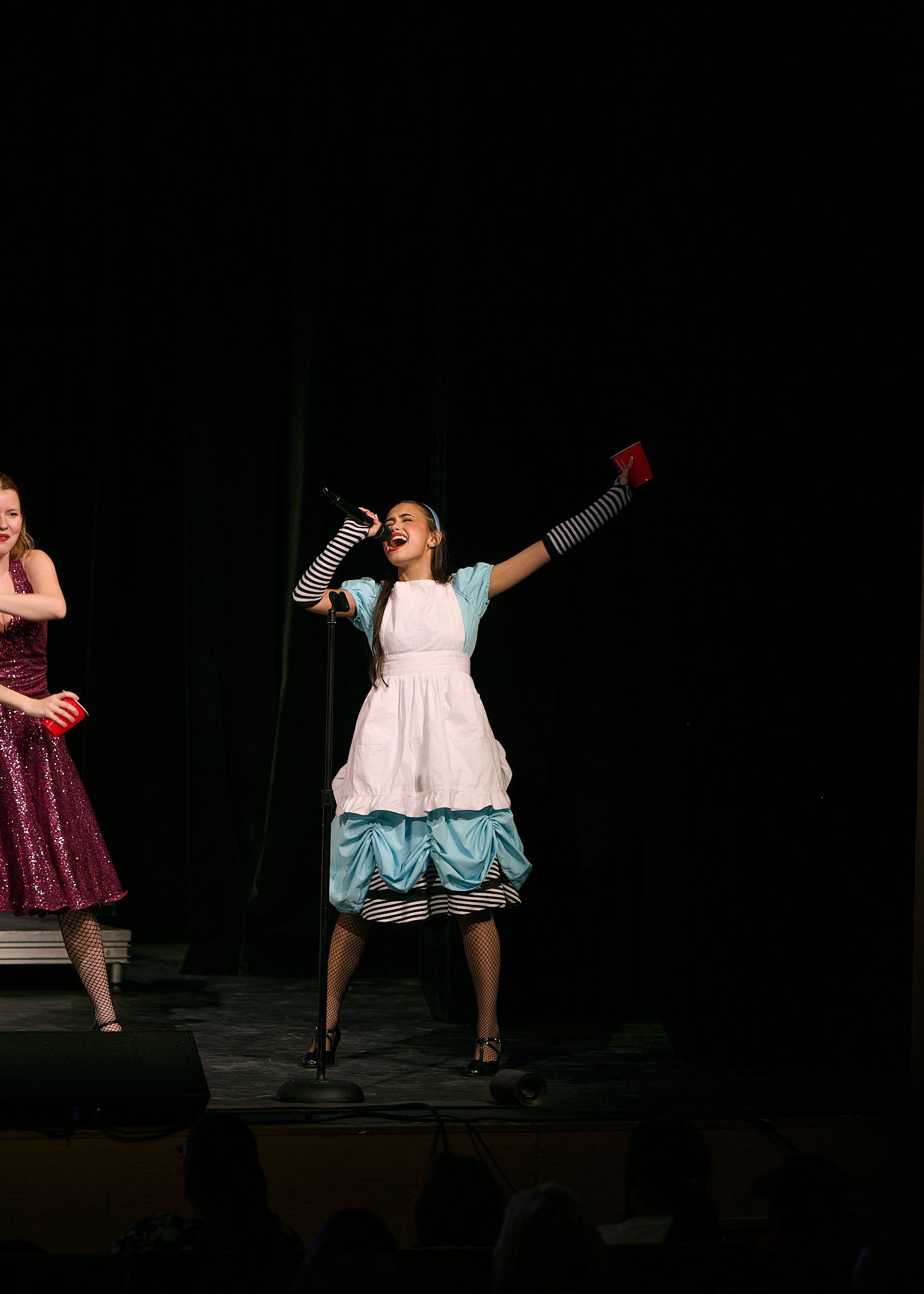 A girl dressed as Alice in Wonderland performs on stage with arms raised during a show choir competition.
