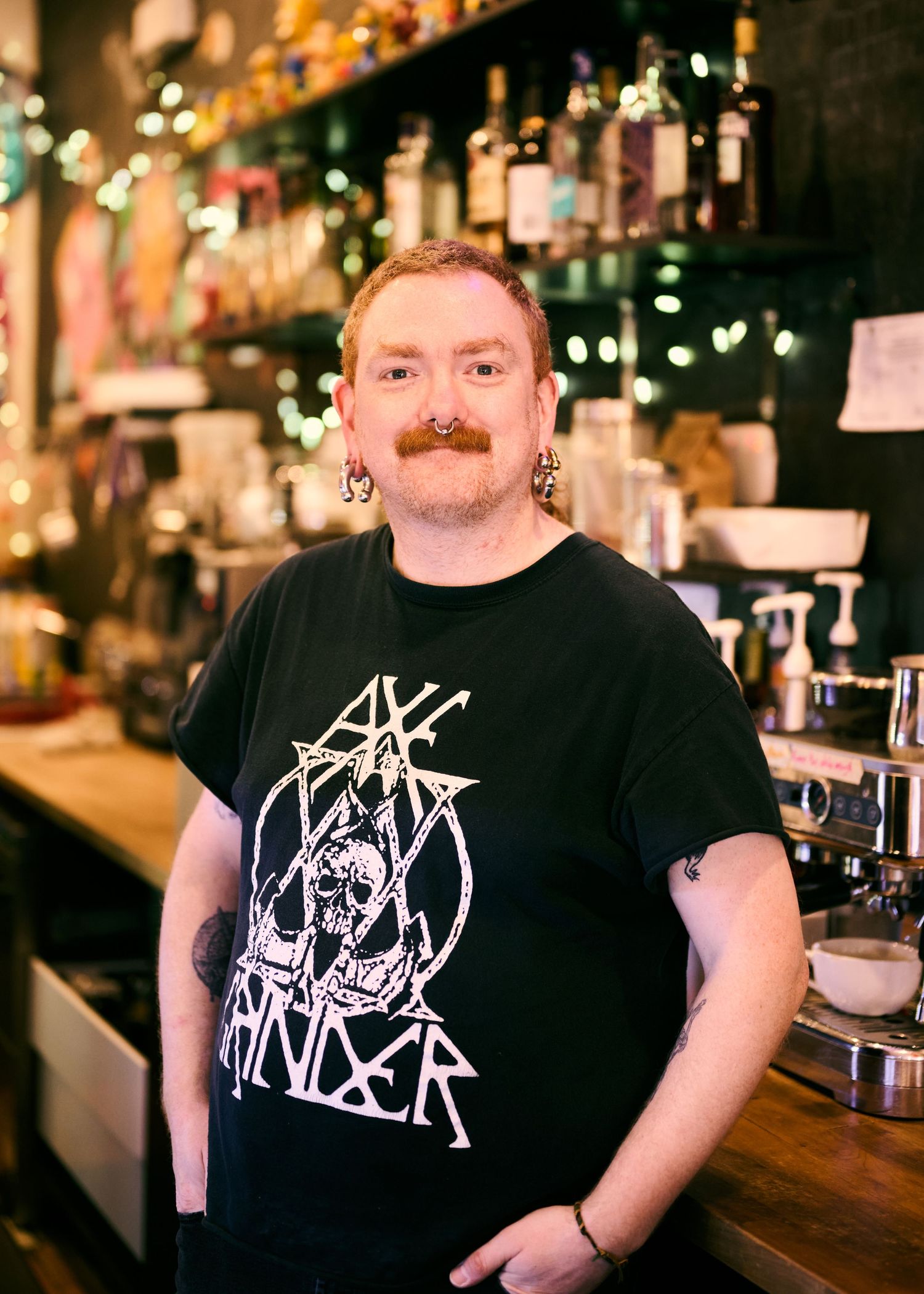 A bartender in a black band t-shirt stands behind a well-stocked bar with liquor bottles visible in the background.
