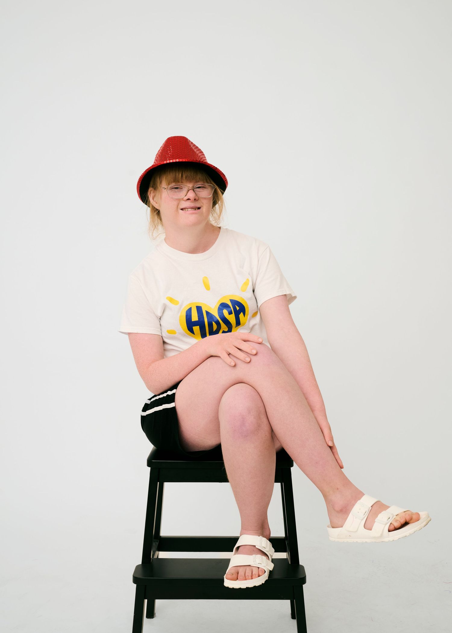 Person in white t-shirt and red hat sits casually on black stool against white background.