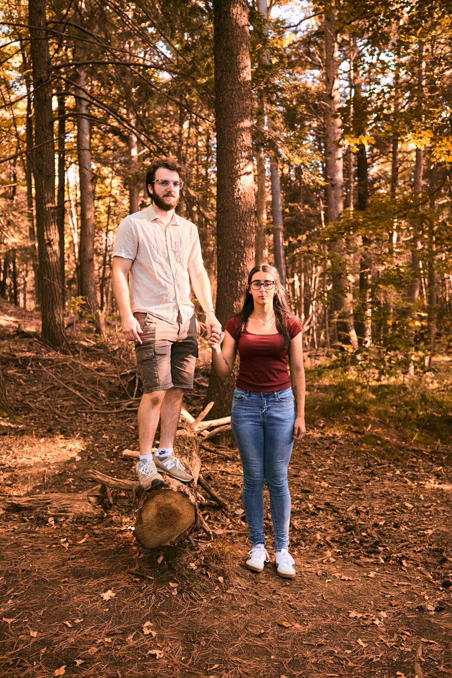 A couple stands together on a wooded trail during golden hour with autumn leaves scattered on the forest floor.