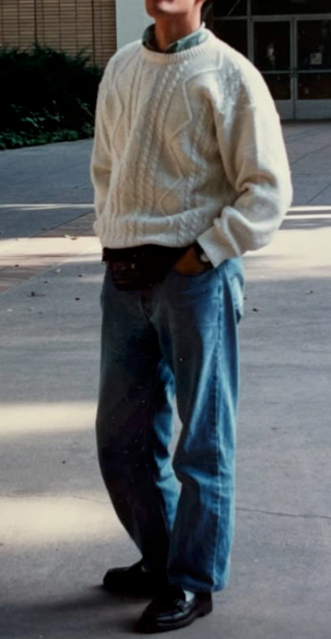 Person wearing a cream cable knit sweater and blue jeans stands on concrete pavement in outdoor setting.