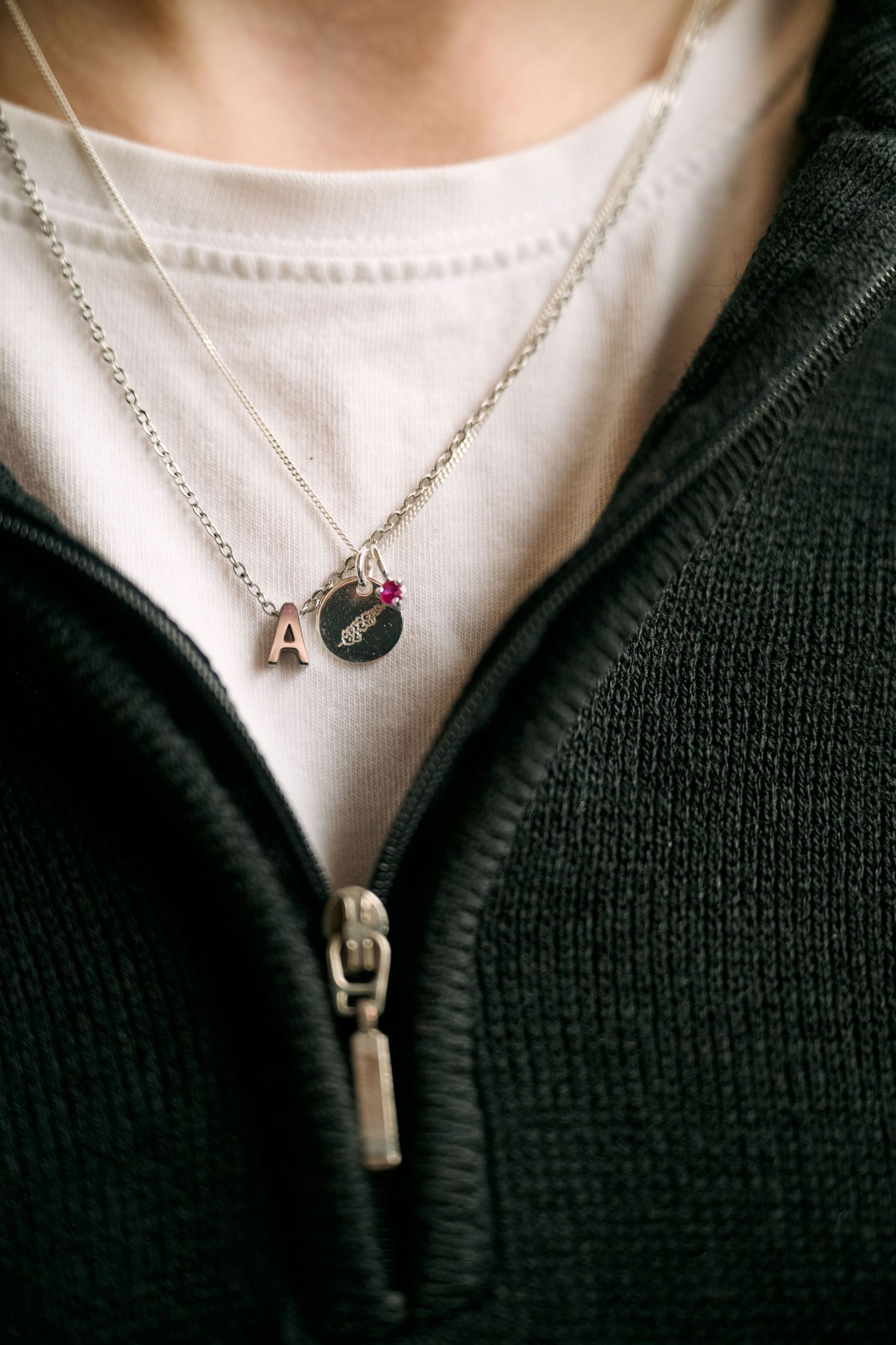 A delicate silver chain necklace with small letter and charm pendants worn over a white shirt and black zippered sweater.