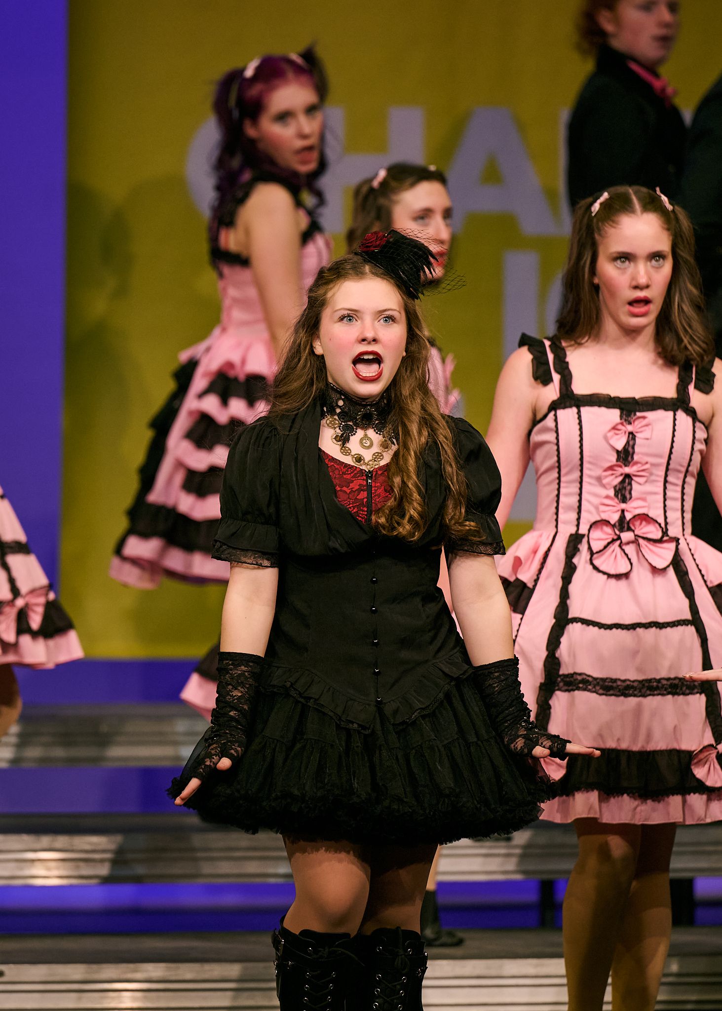 Young performers in black and pink costumes sing on stage during a theatrical production.