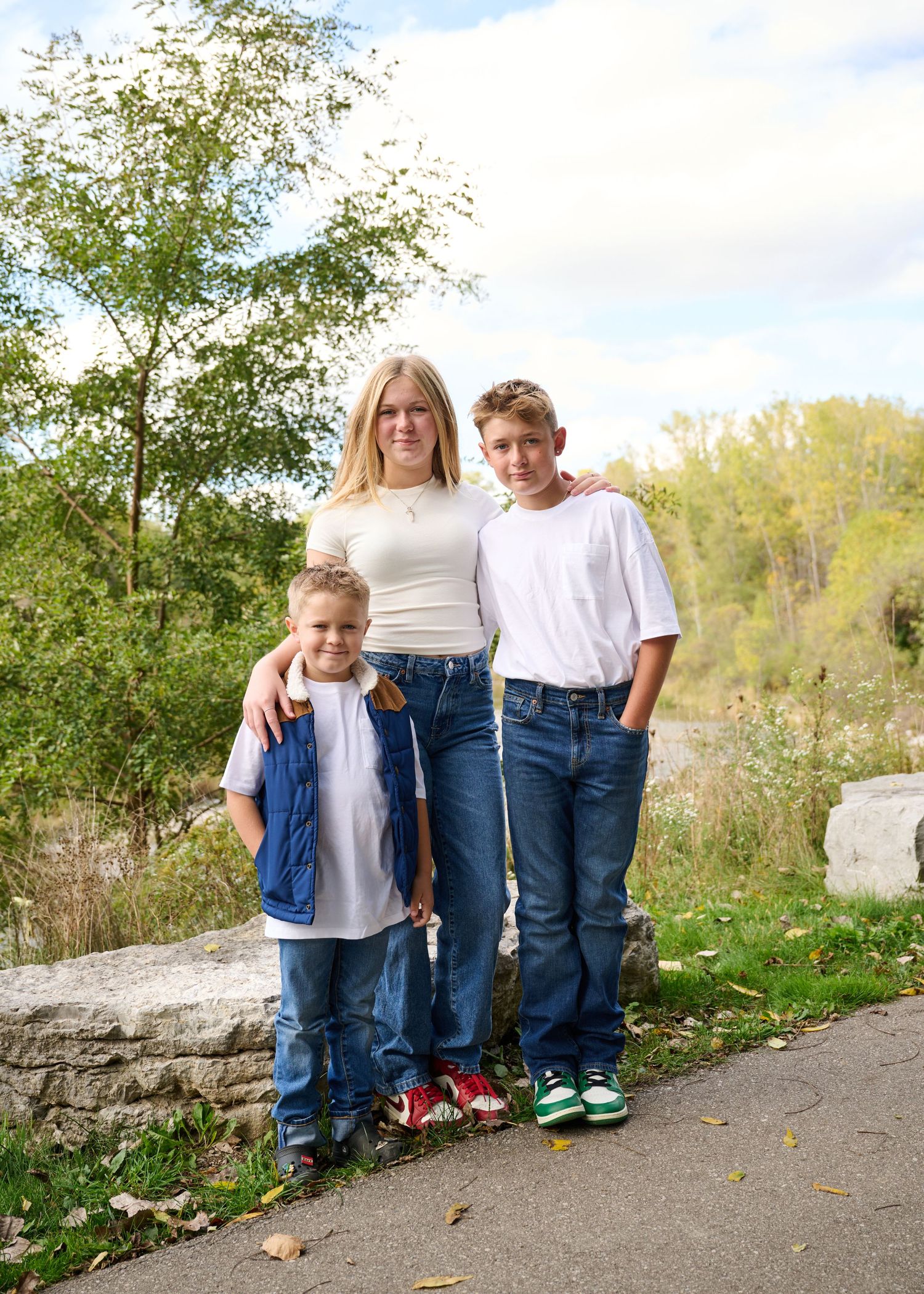 Three people stand together on a stone path outdoors with trees and greenery in the background on a sunny day.