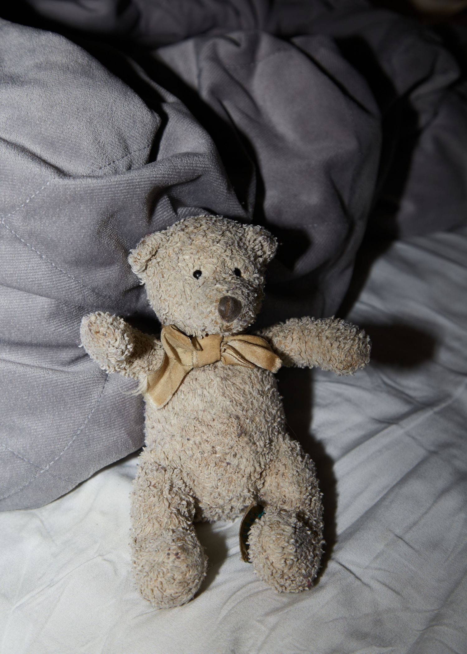 A plush teddy bear with a beige bow sitting on gray bedding.