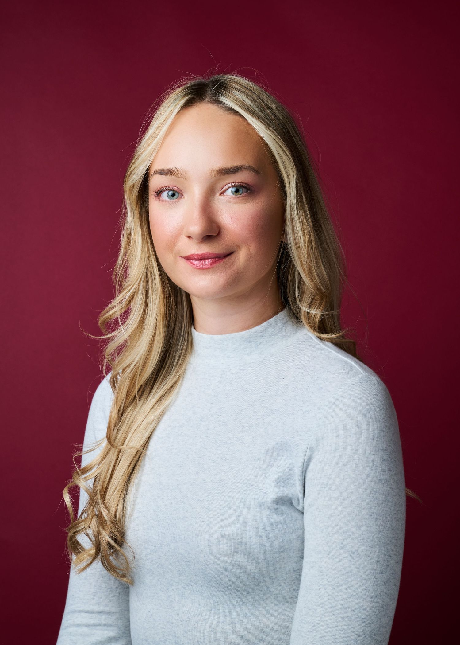 Professional headshot of a blonde person in a light gray sweater against a burgundy background.