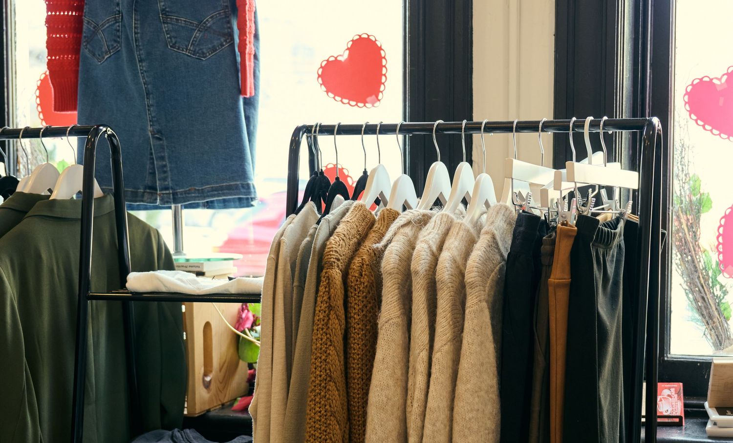 Clothing rack displaying brown sweaters and jeans with heart decorations on window in retail store setting.