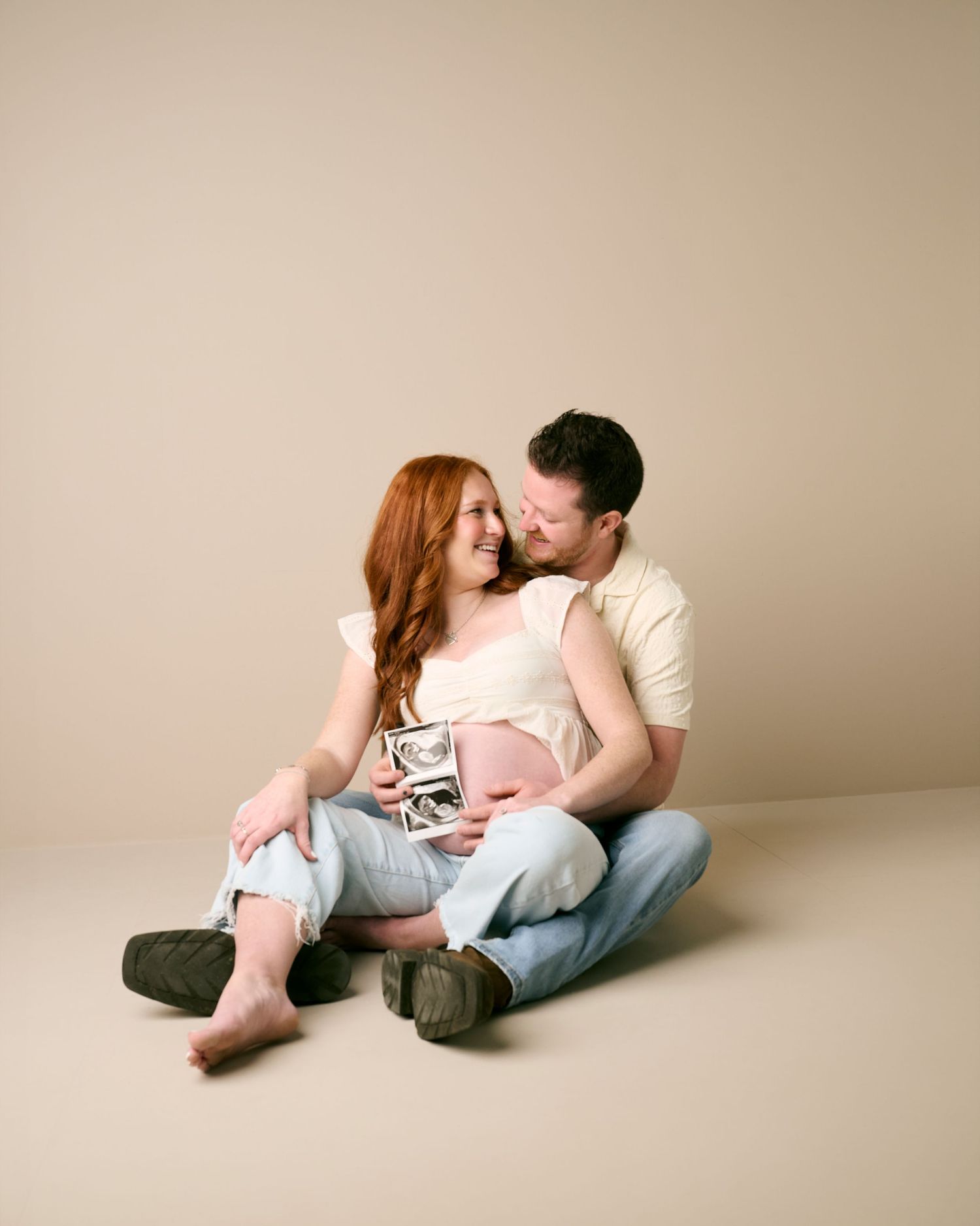 A couple in casual attire sitting close together on the floor in a minimalist beige studio setting.