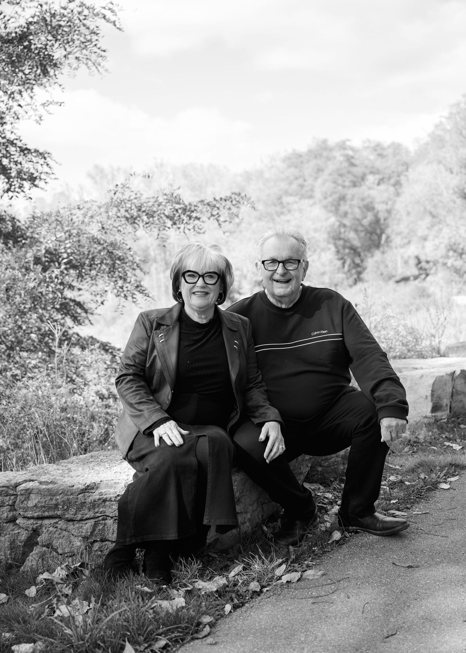 A couple sits together on a stone wall in a black and white photograph outdoors.