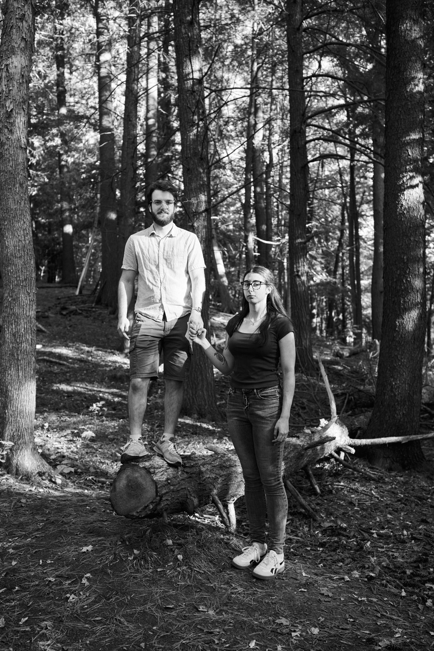 Black and white photo of a young couple standing on a fallen log in a forest setting.