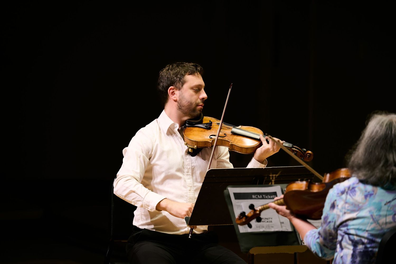 Male violinist performing on stage with a music stand, accompanied by another musician in a concert setting.