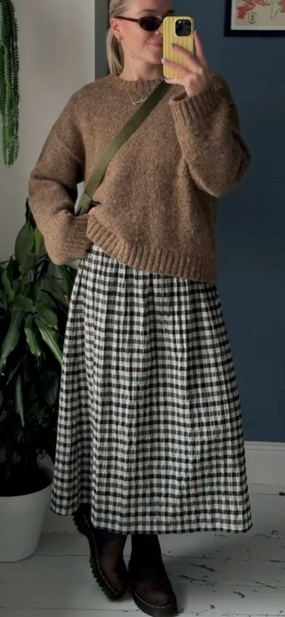 A cozy fall outfit featuring a brown knit sweater paired with a black and white gingham midi skirt and dark boots.