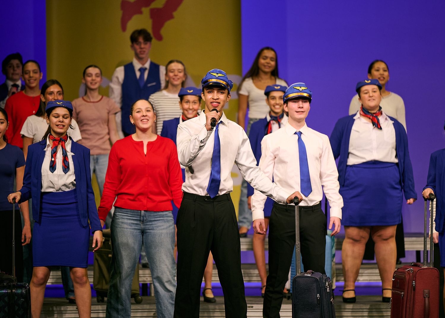 Students perform a theatrical production in airline crew costumes, with rolling luggage on a colorful stage set.