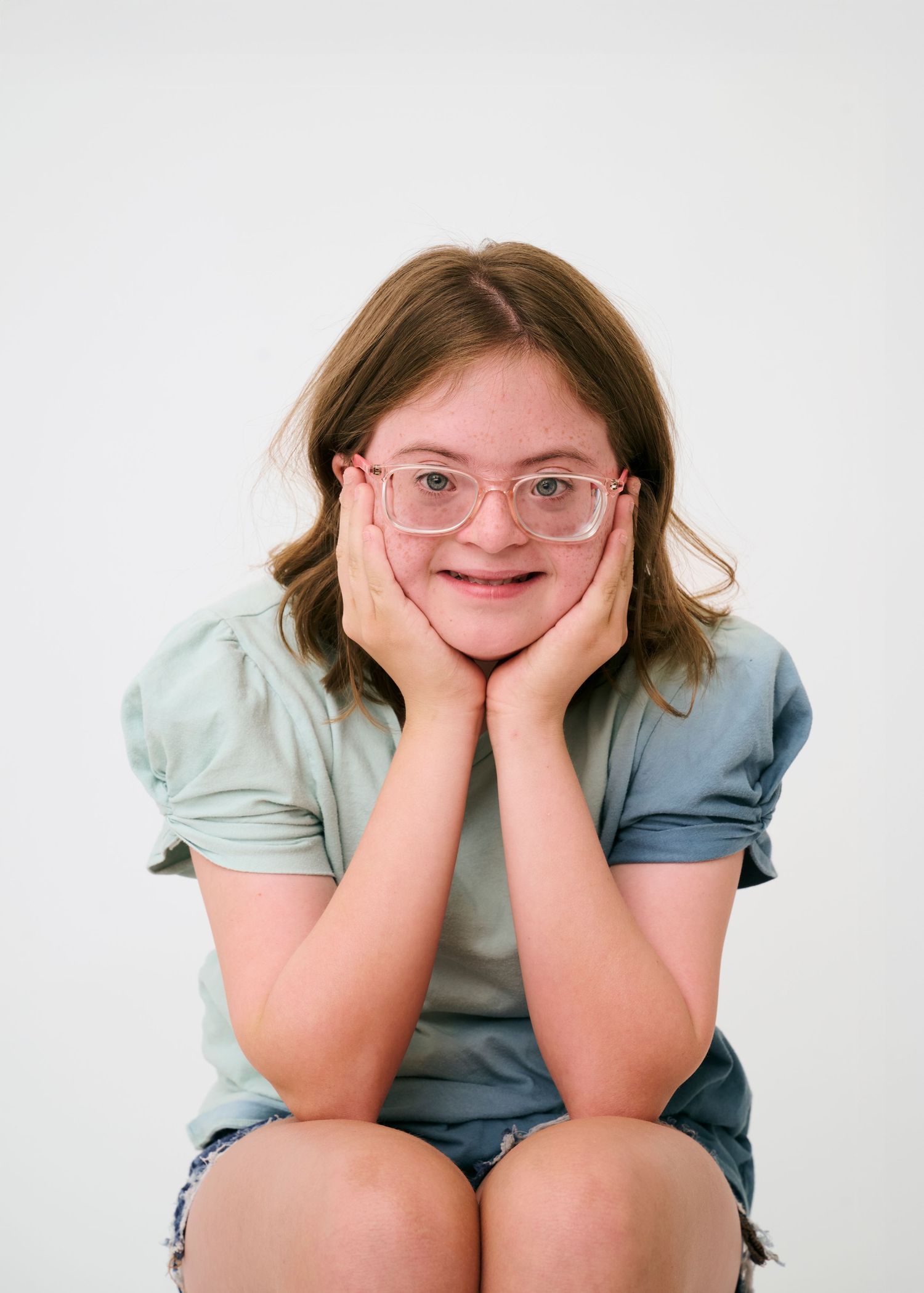 Person wearing glasses and light blue shirt poses playfully with hands on cheeks against white background.