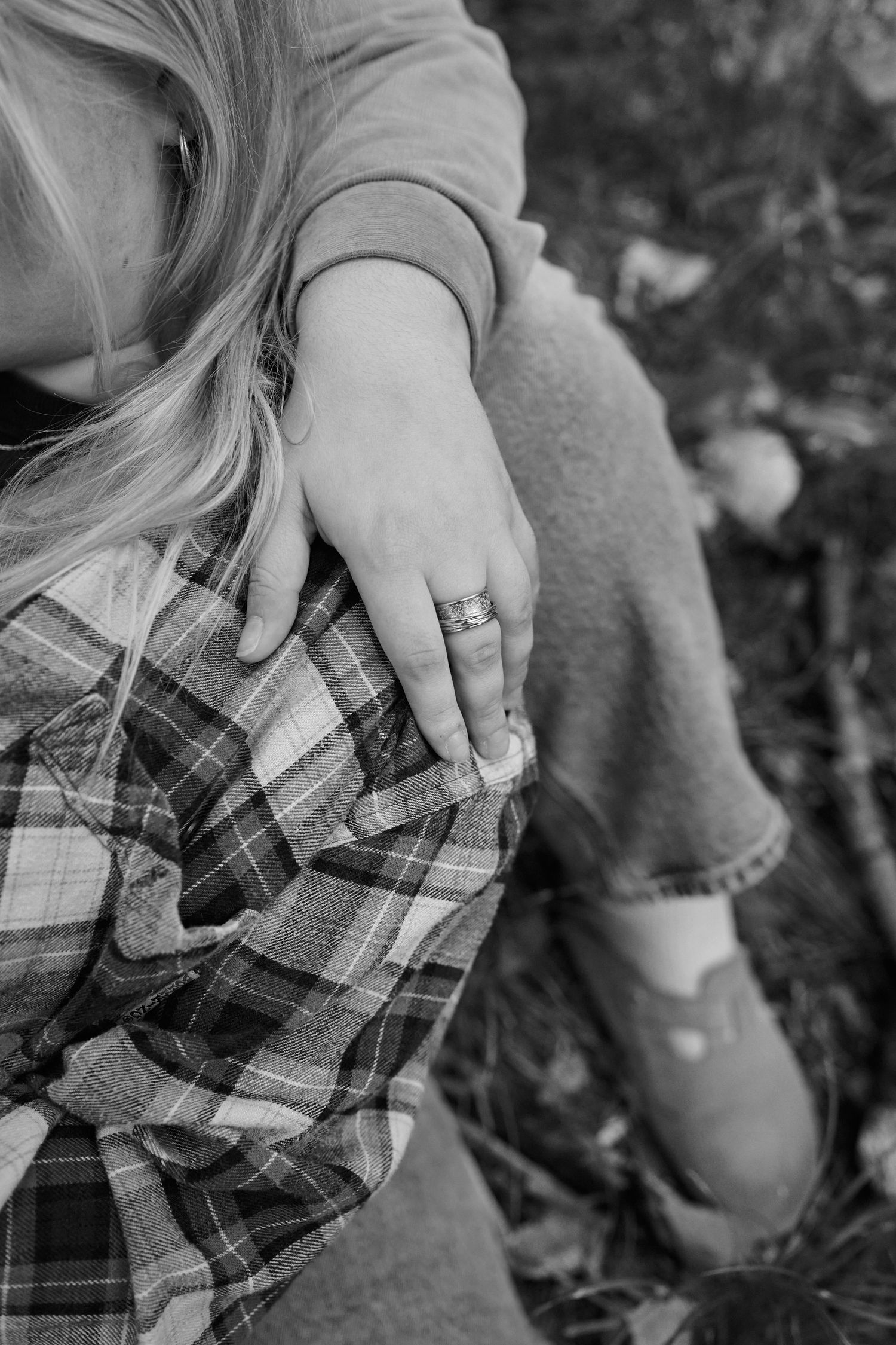 A close-up black and white photo shows hands wearing a ring resting on a plaid shirt sleeve.