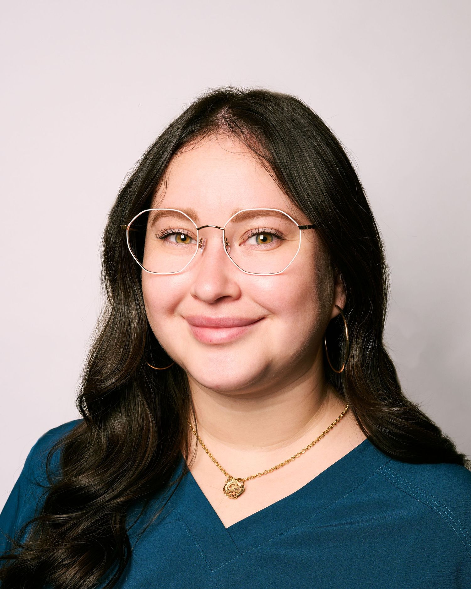Young woman with dark wavy hair, round gold glasses, hoop earrings, gold necklace, wearing a teal scrub top.