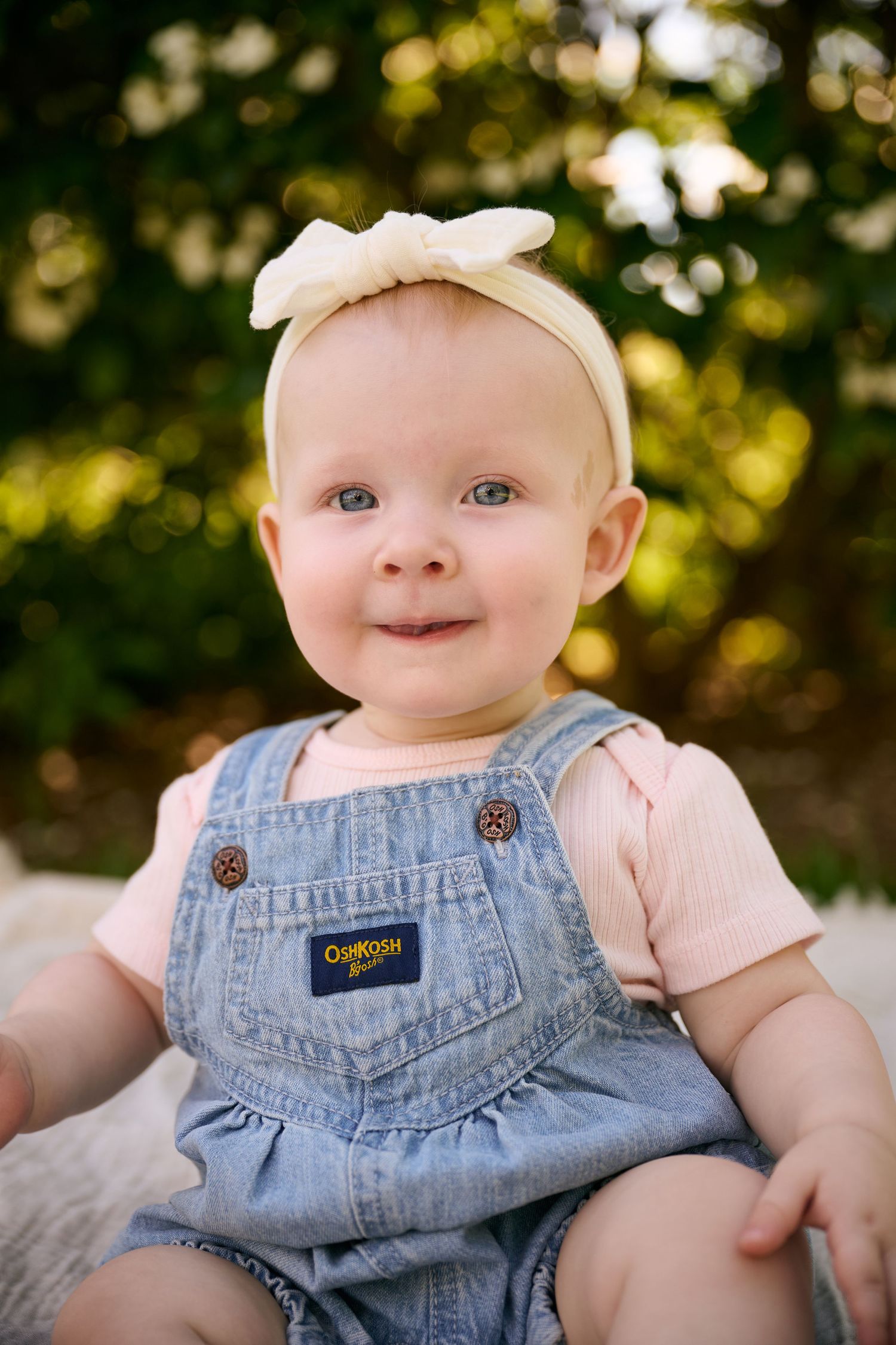 Smiling baby girl in denim OshKosh overalls and cream bow headband sitting outdoors with green trees behind her.