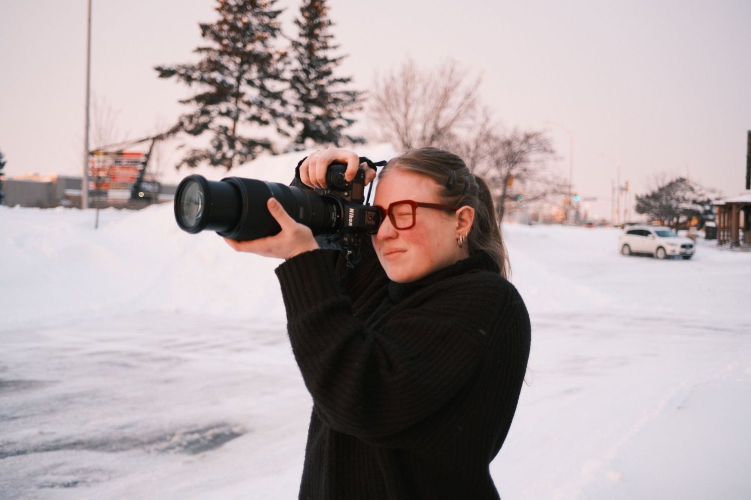 A photographer wearing a black sweater uses a telephoto lens to capture shots in a snowy winter landscape.