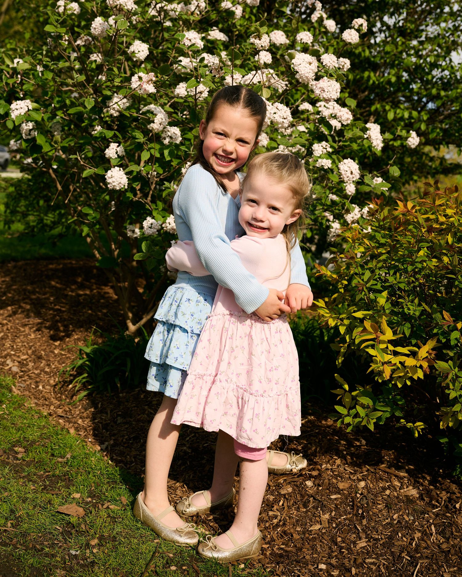 Two young girls hugging and smiling in front of a flowering bush, wearing pastel dresses outdoors.