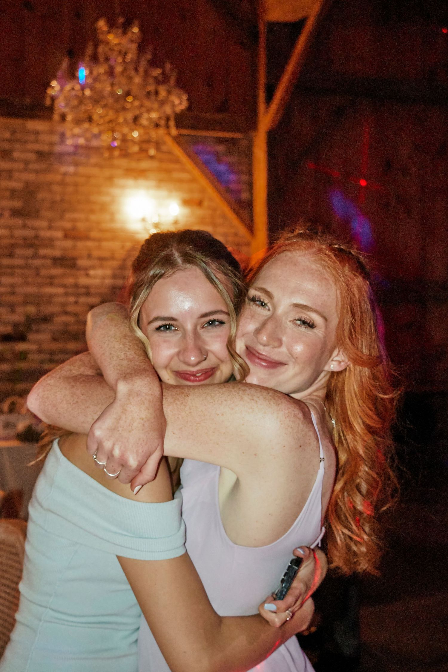 Two friends embrace and smile warmly while dancing under a chandelier in a dimly lit venue.