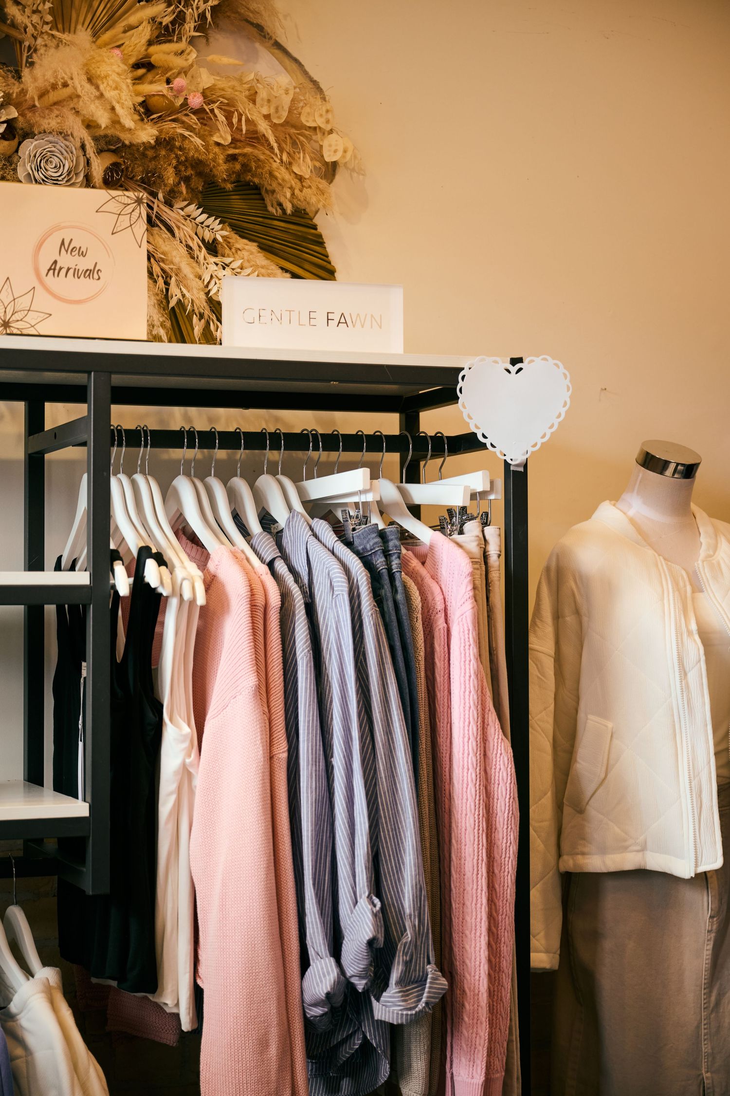 A clothing rack displays pastel and neutral colored garments in a boutique with a white mannequin and decorative details.