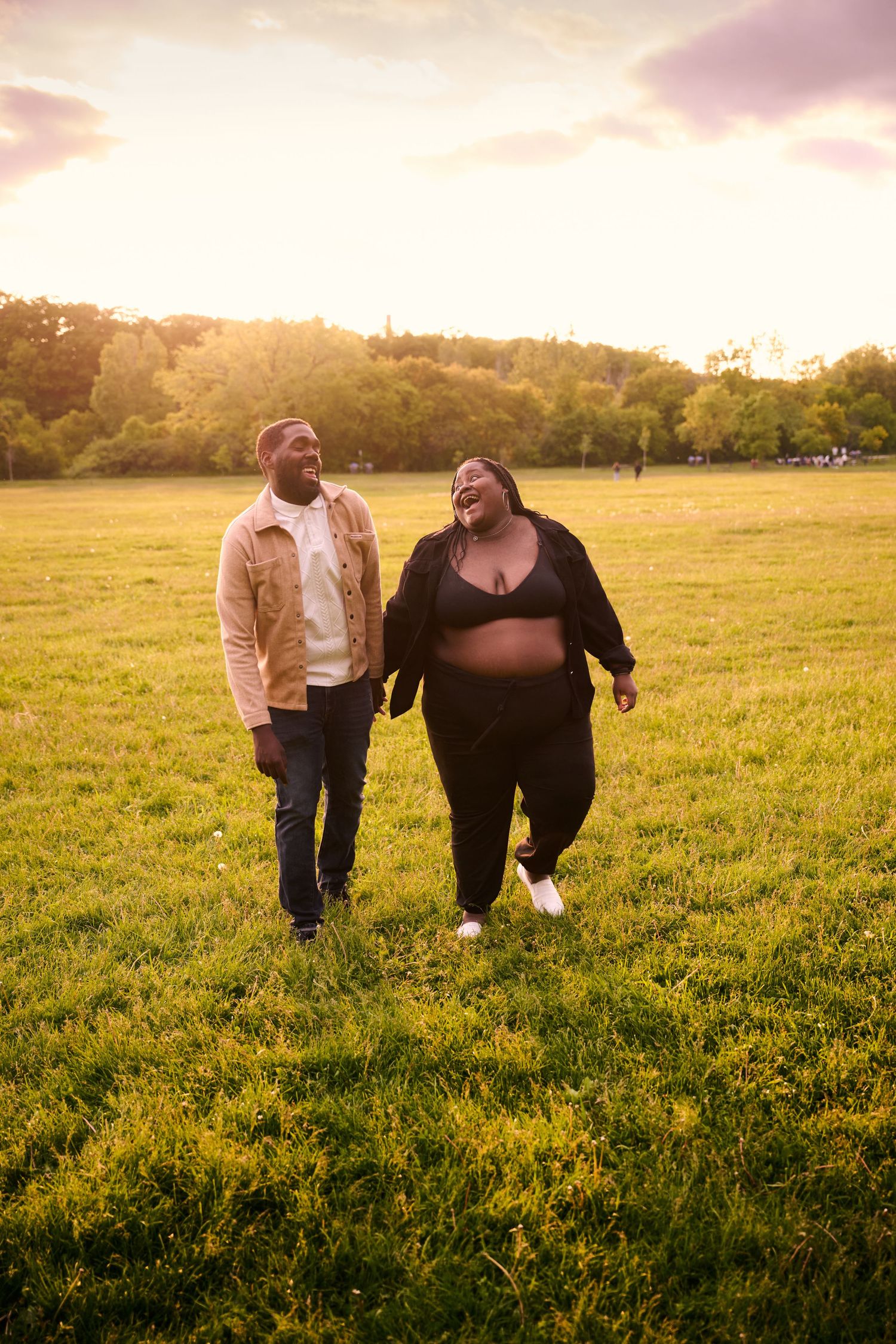 A couple walks together through a sunlit meadow at golden hour as warm light bathes the grassy field.