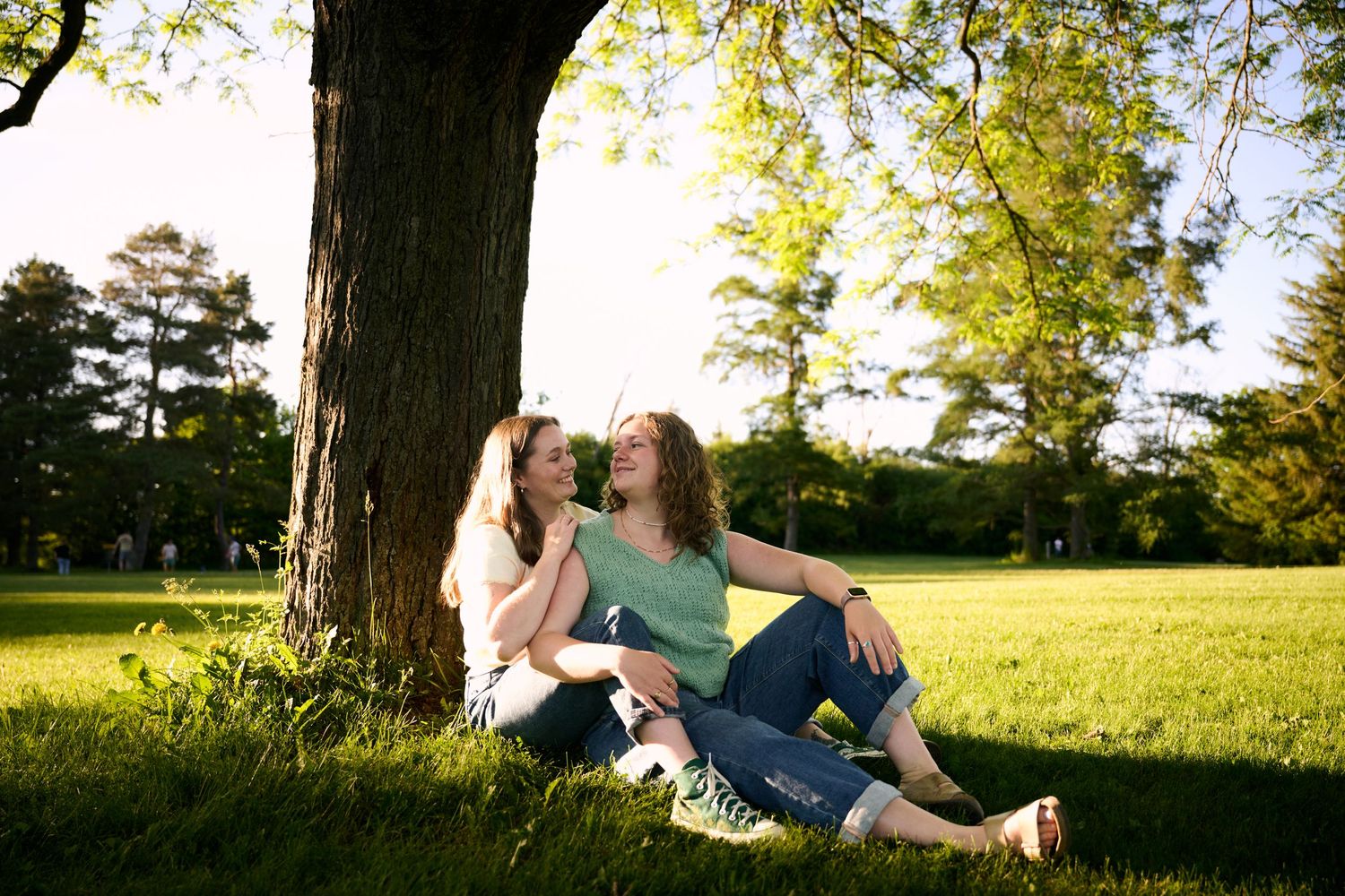 Two women sitting together under a large tree in a sunny green park, smiling and enjoying each other's company.