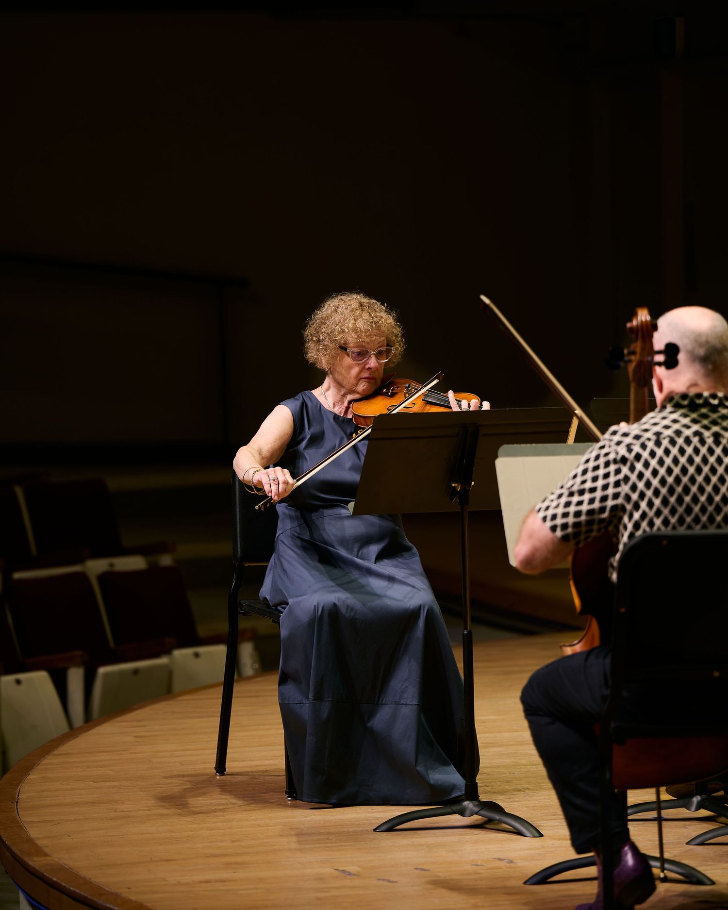 Woman in navy dress plays violin on stage while seated, performing chamber music with another musician in a concert hall.