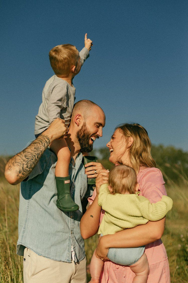 Happy family with tattooed dad, mom, toddler on shoulders, and baby enjoying sunny outdoor field day.