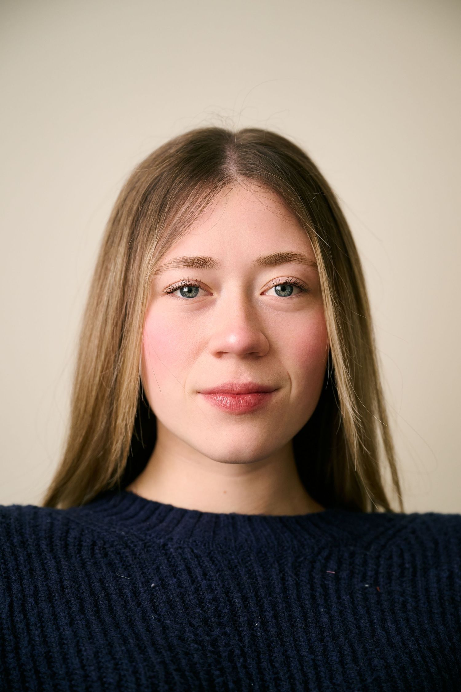 Young woman with long blonde hair and green eyes wearing a navy knit sweater against a neutral background.
