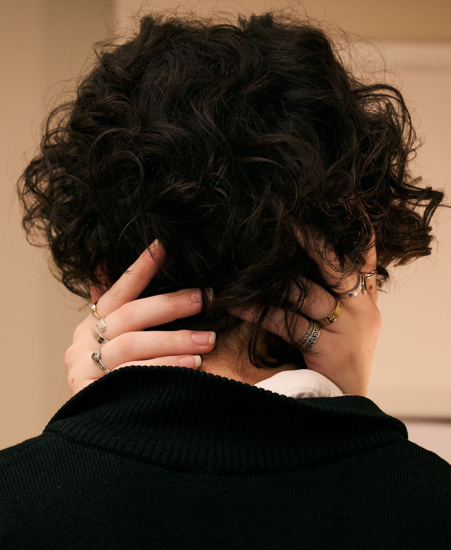 View from behind of dark curly hair with a hand wearing rings running through it against a beige background.