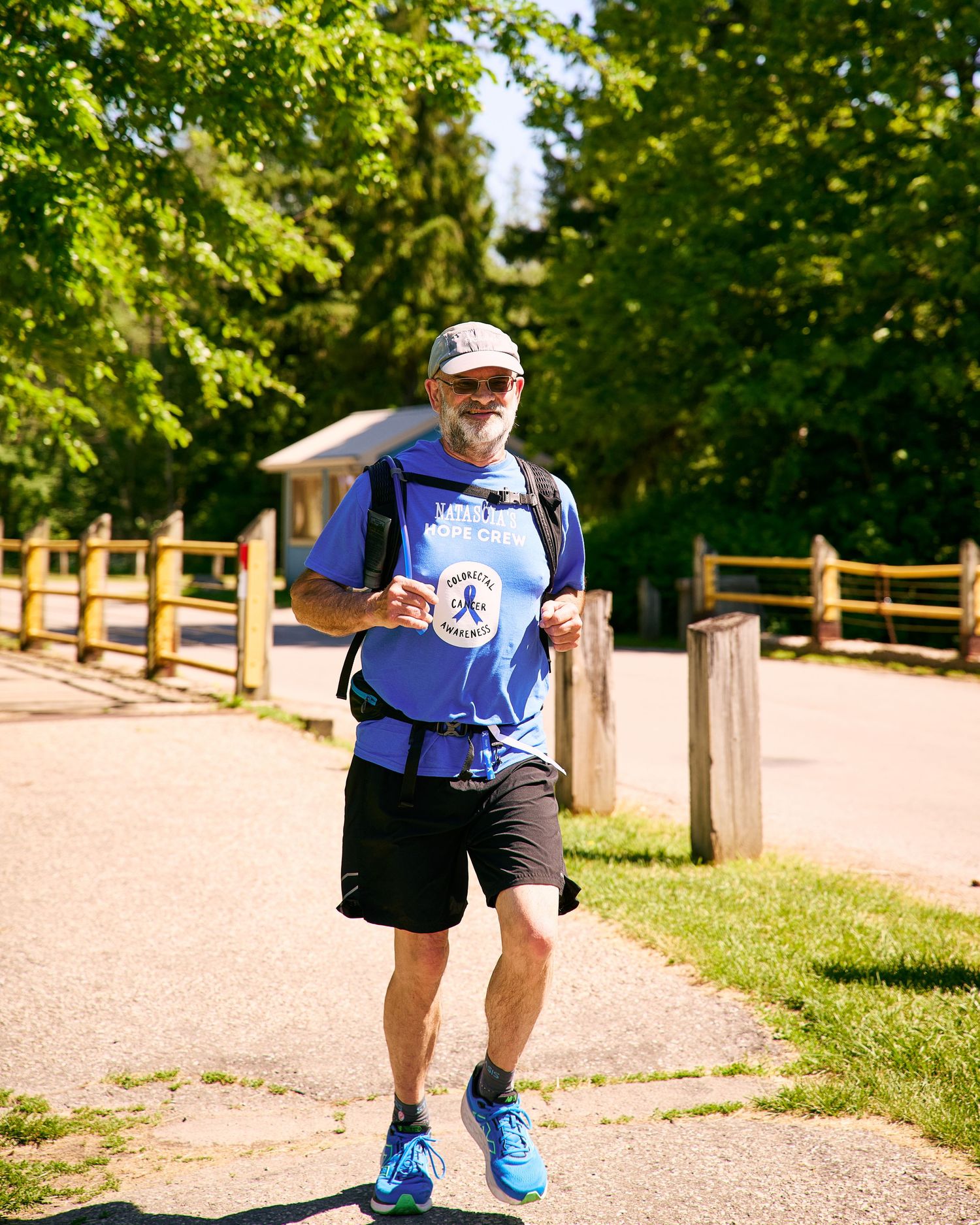Older man in blue shirt and cap jogging on a sunny paved path surrounded by green trees and wooden fence.