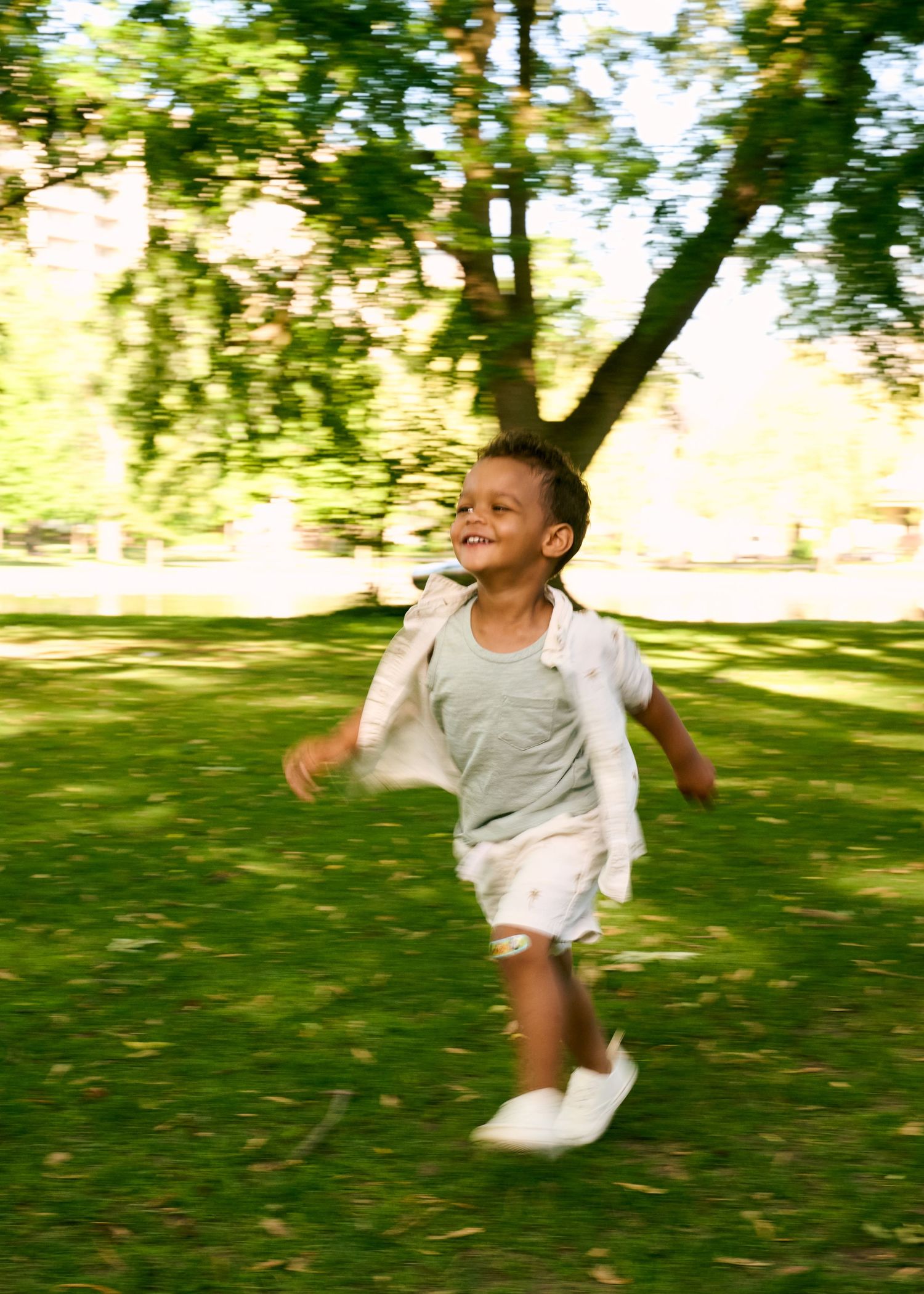A joyful young boy runs through a sunny green park, wearing a gray shirt, white shorts, and white sneakers.