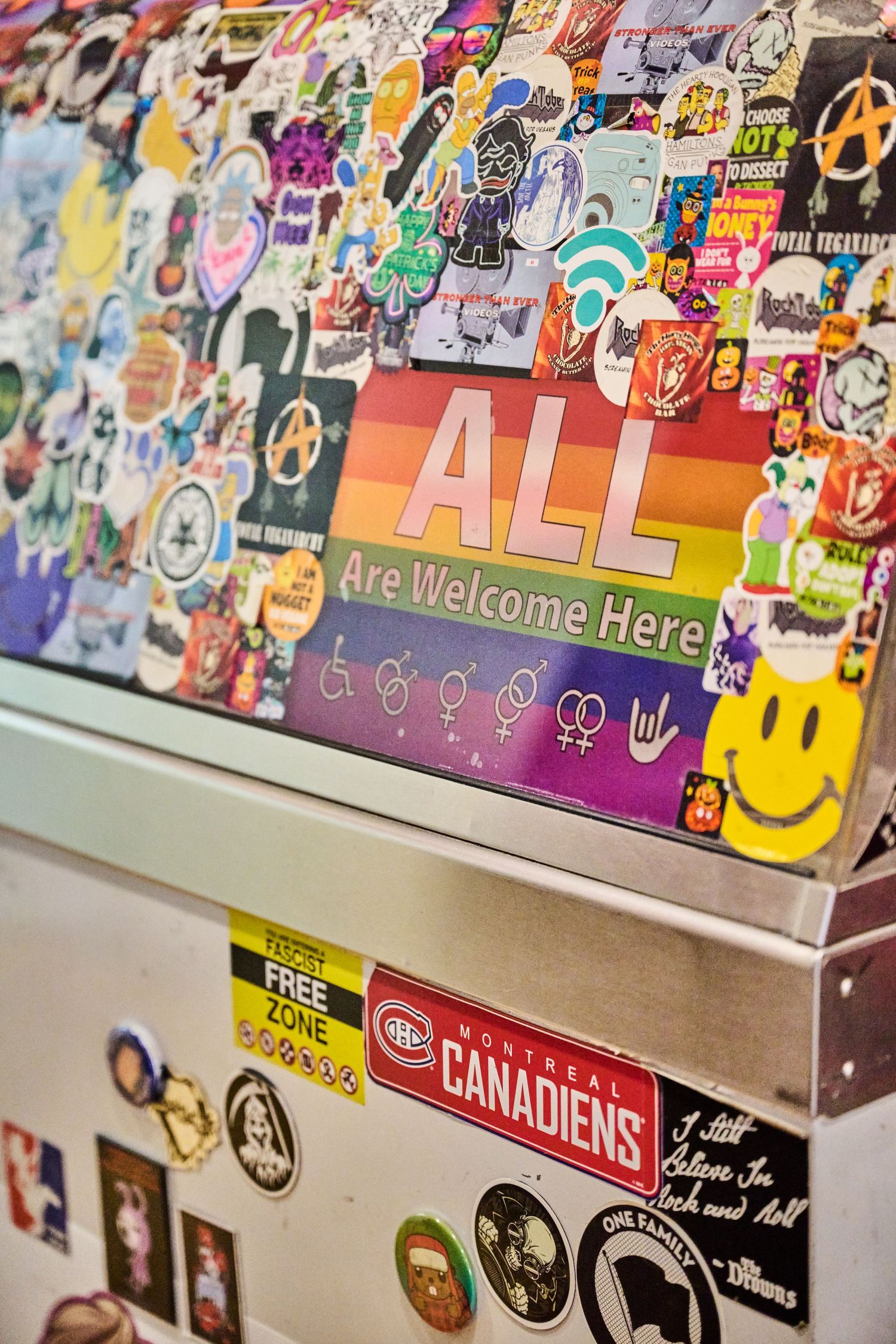 A laptop covered in colorful stickers including a rainbow 'ALL Are Welcome Here' message and sports team logos.