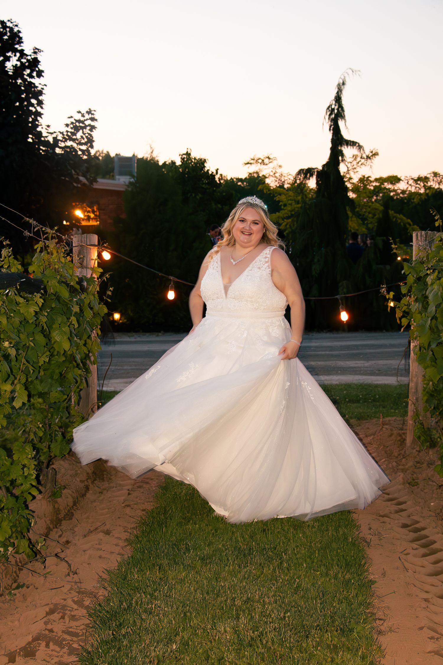 An ethereal figure in a white tulle gown twirls gracefully among vineyard rows at sunset with string lights overhead.