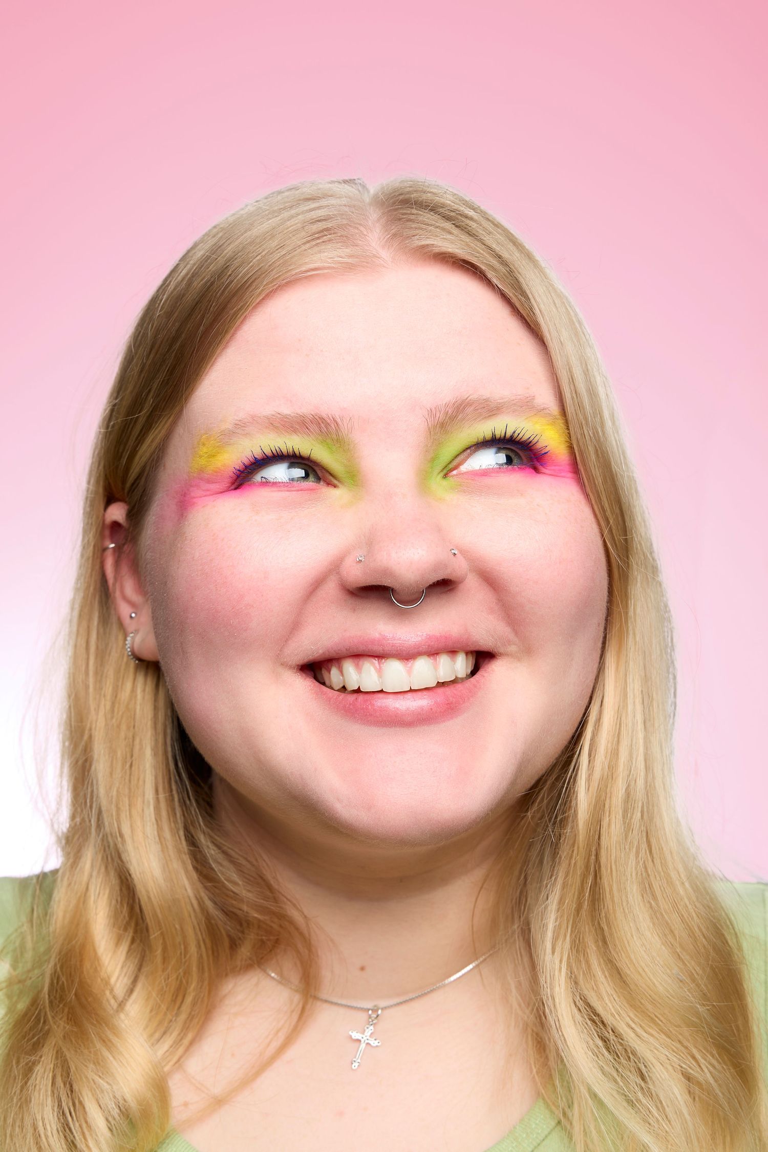 Portrait against pink background showing bright yellow and green eye makeup with long blonde hair and joyful expression.
