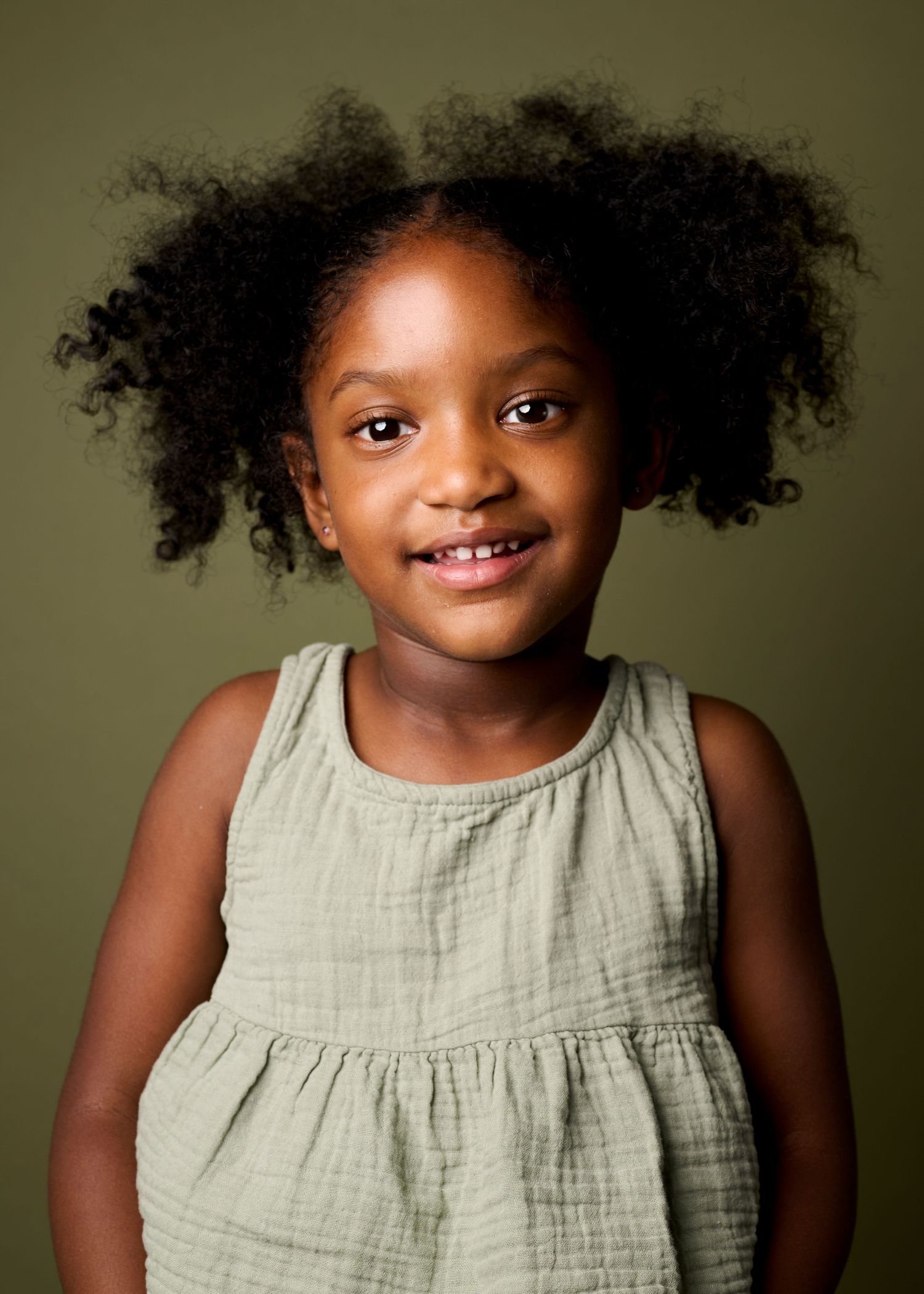 A young child with natural curly hair wearing a sage green dress smiles against an olive background.