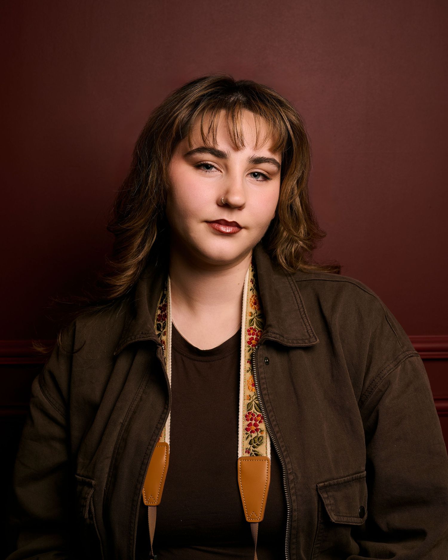 Portrait against dark brown background wearing brown jacket with floral pattern lining and dark top.